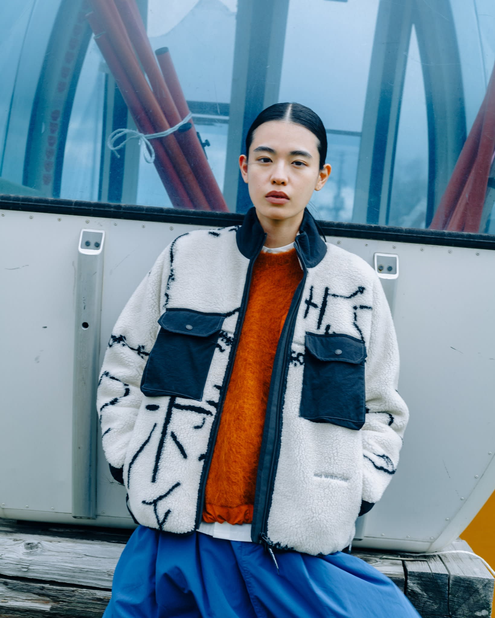 A young woman in a patterned fleece jacket and orange sweater poses in front of a metallic structure with red pipes.