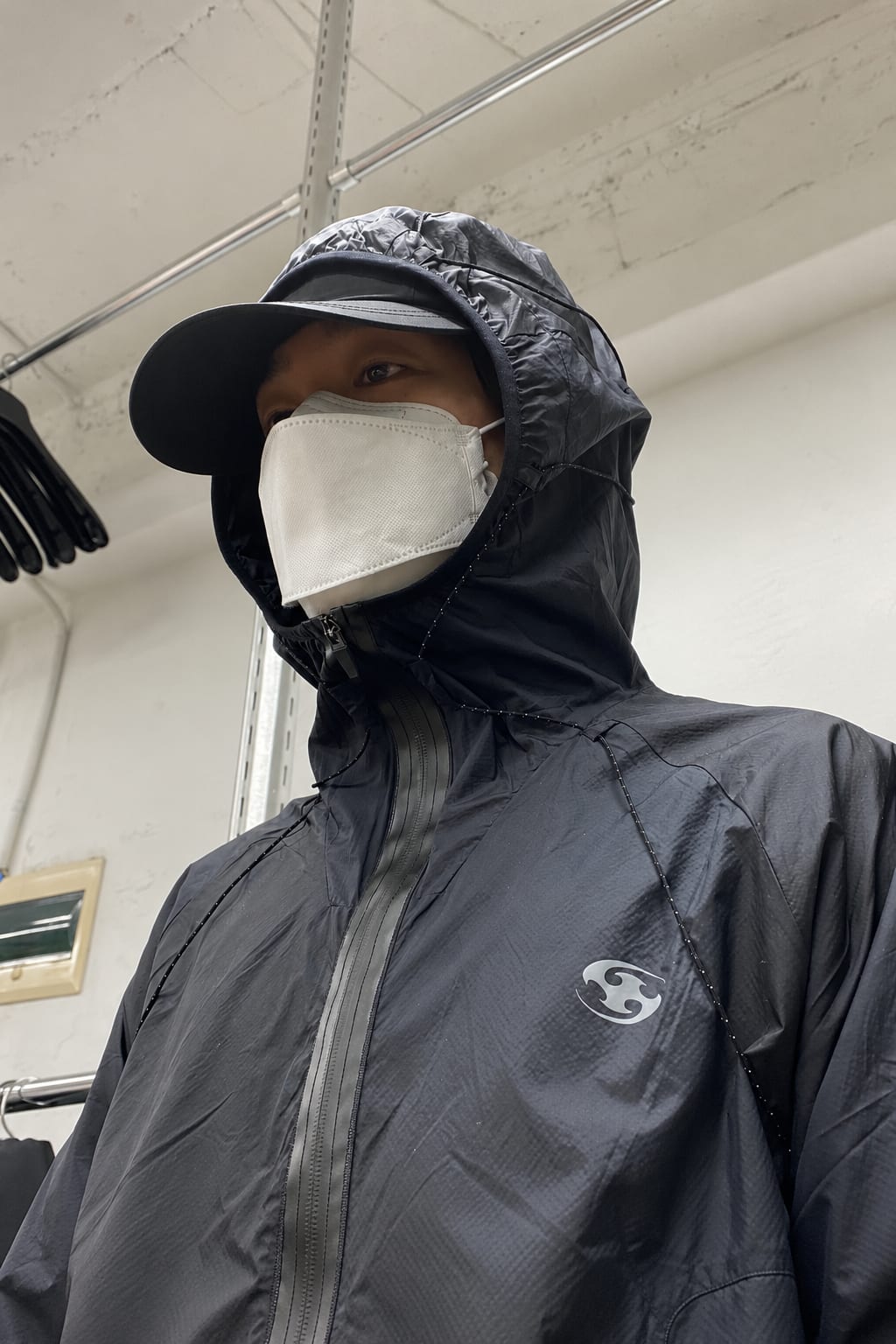 A person wearing a black hooded jacket, a black cap, and a white face mask stands against a plain white wall with metal shelving.