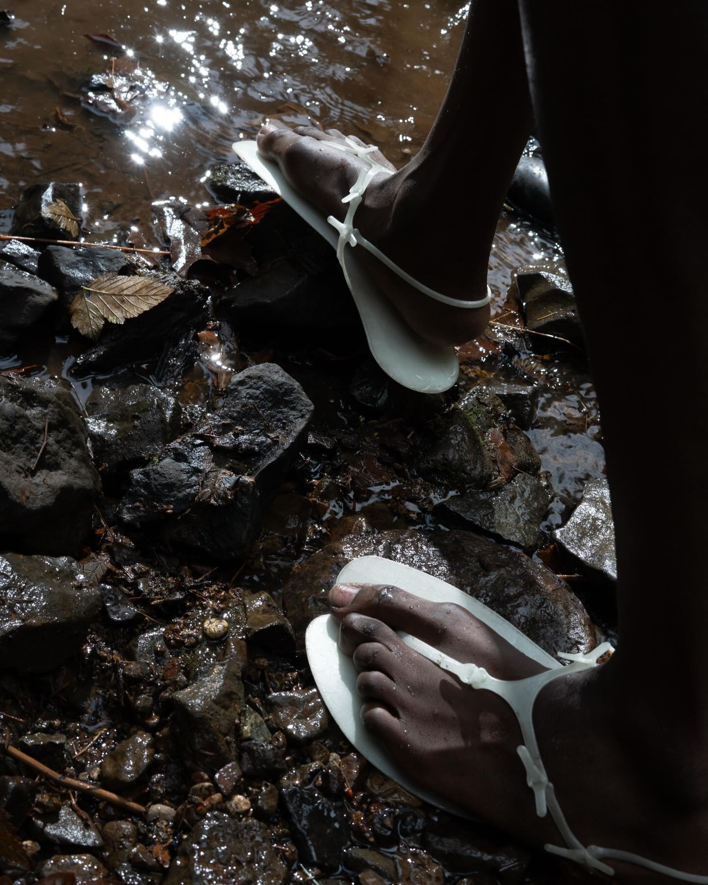 A person wearing white flip-flops stands on dark, wet rocks beside a shimmering body of water.