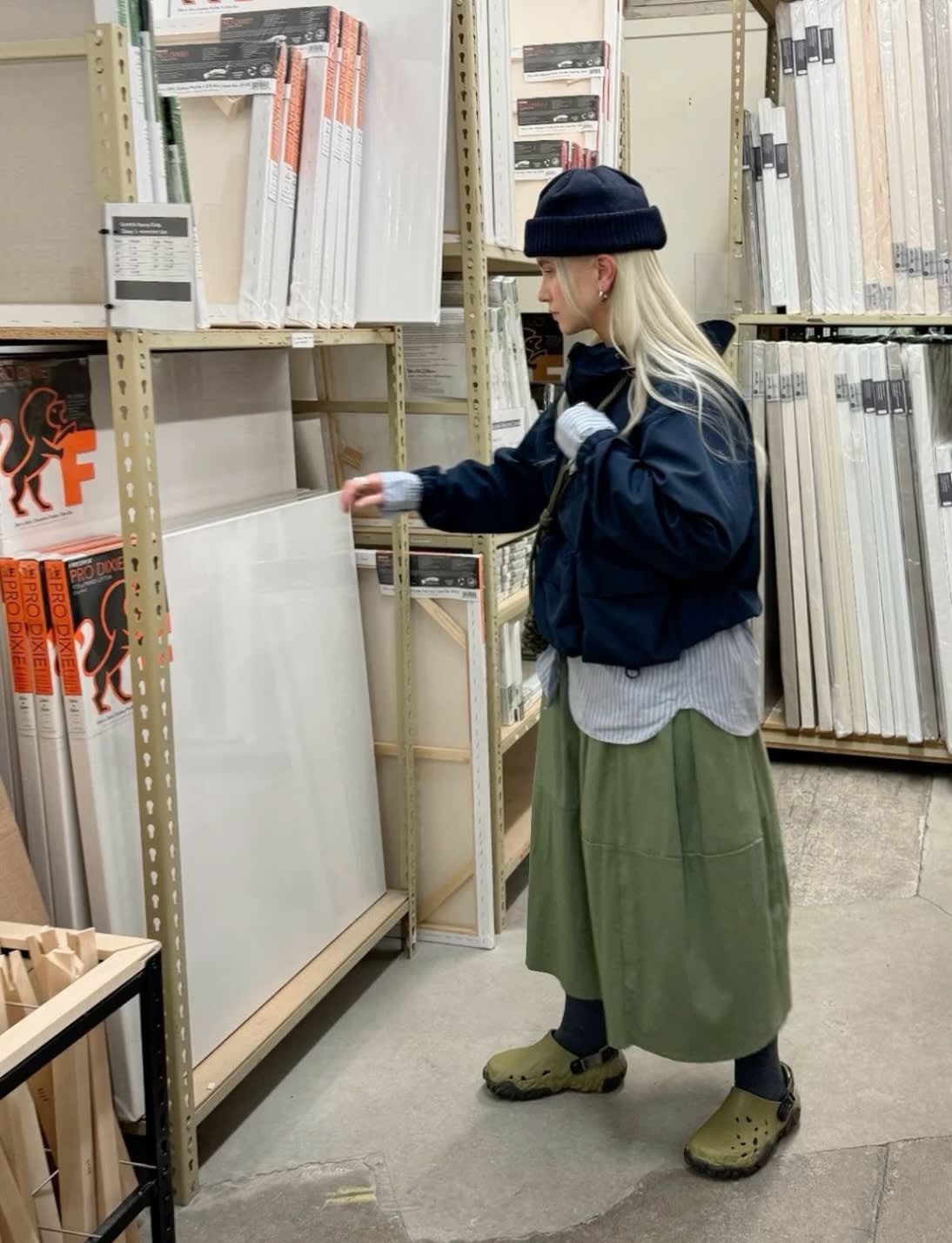 A person in a beanie and green skirt examines a blank canvas on a shelf in an art supply store, surrounded by stacks of canvases and art supplies.