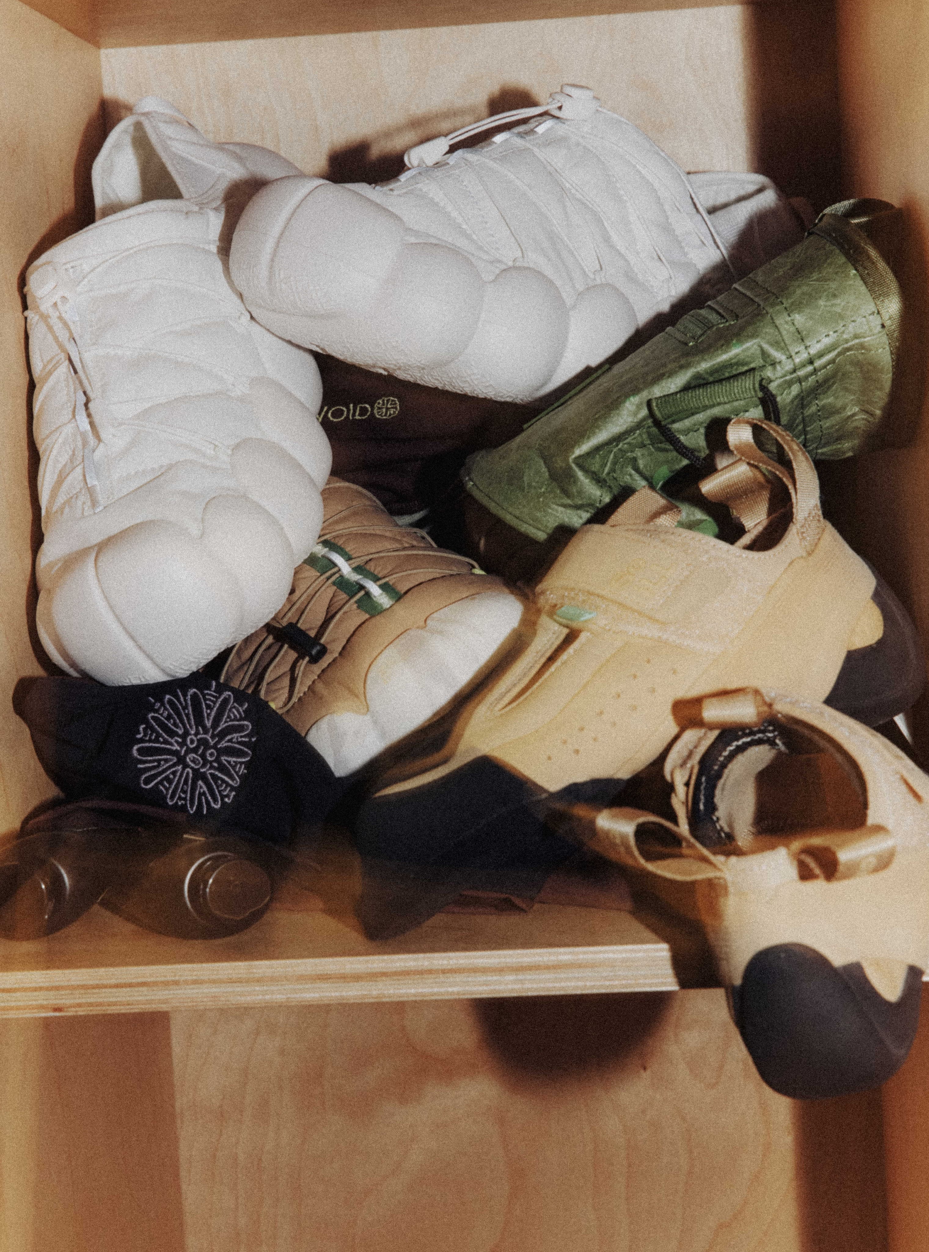 Various climbing shoes in white, green, and tan colors are piled together inside a light-colored wooden shelf.