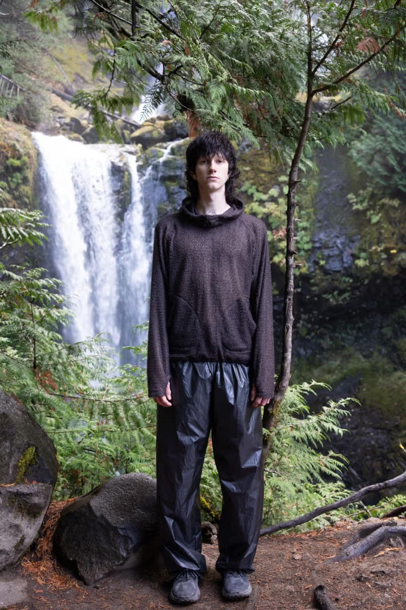 A young man in a brown textured hoodie and black waterproof pants stands in the foreground before a lush forest and a cascading waterfall in the background.