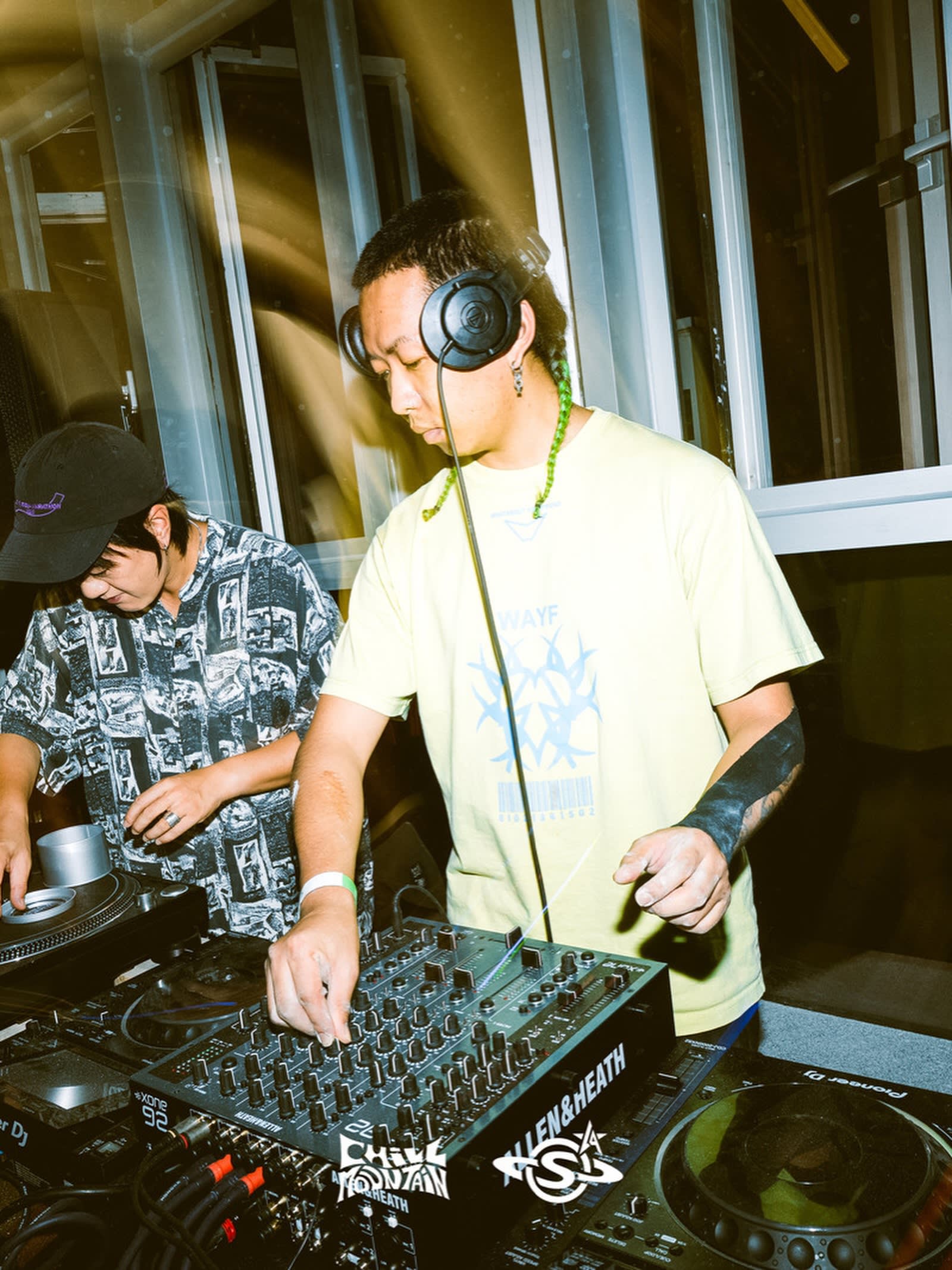 A DJ in a yellow t-shirt operates a mixer in the foreground, while another person in a patterned shirt stands beside him against a background of large windows.