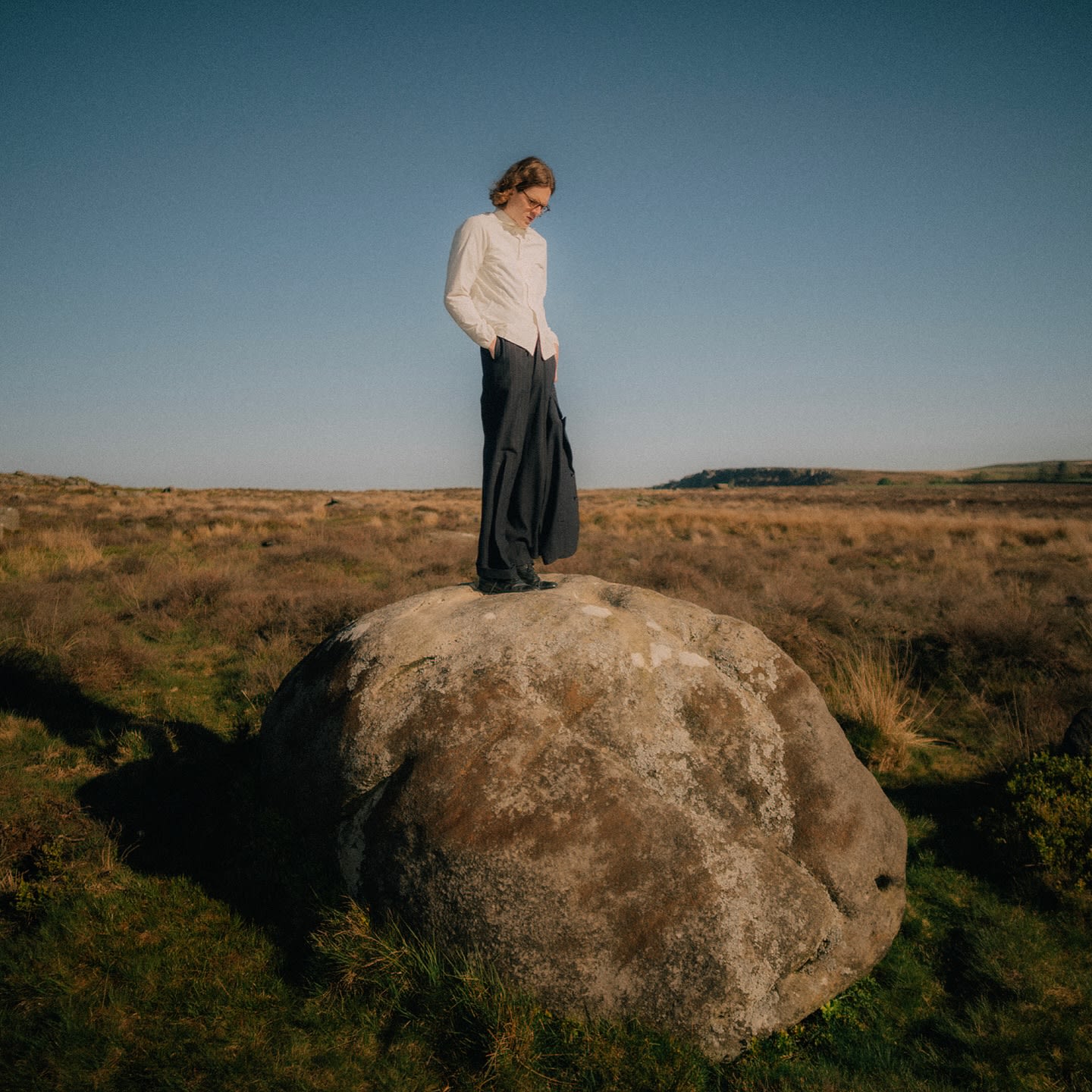 A person in a white shirt and dark trousers stands atop a large boulder in a vast, grassy moorland under a clear blue sky.