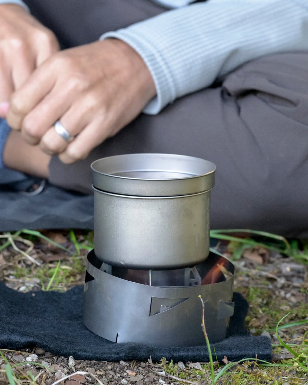 A metal cooking pot sits atop a small camping stove with a fire burning inside, while a person rests in the blurred background.