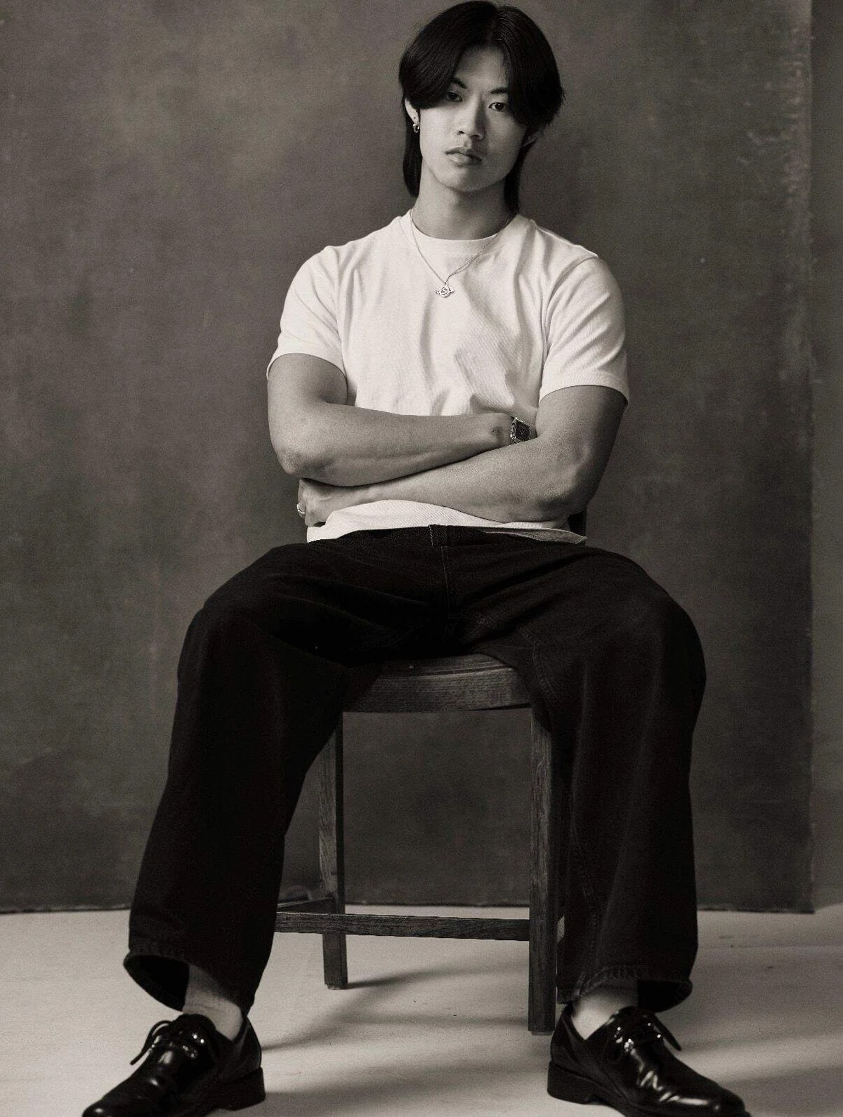 A young man in a white t-shirt and dark trousers sits with crossed arms on a wooden chair against a plain, textured grey wall.