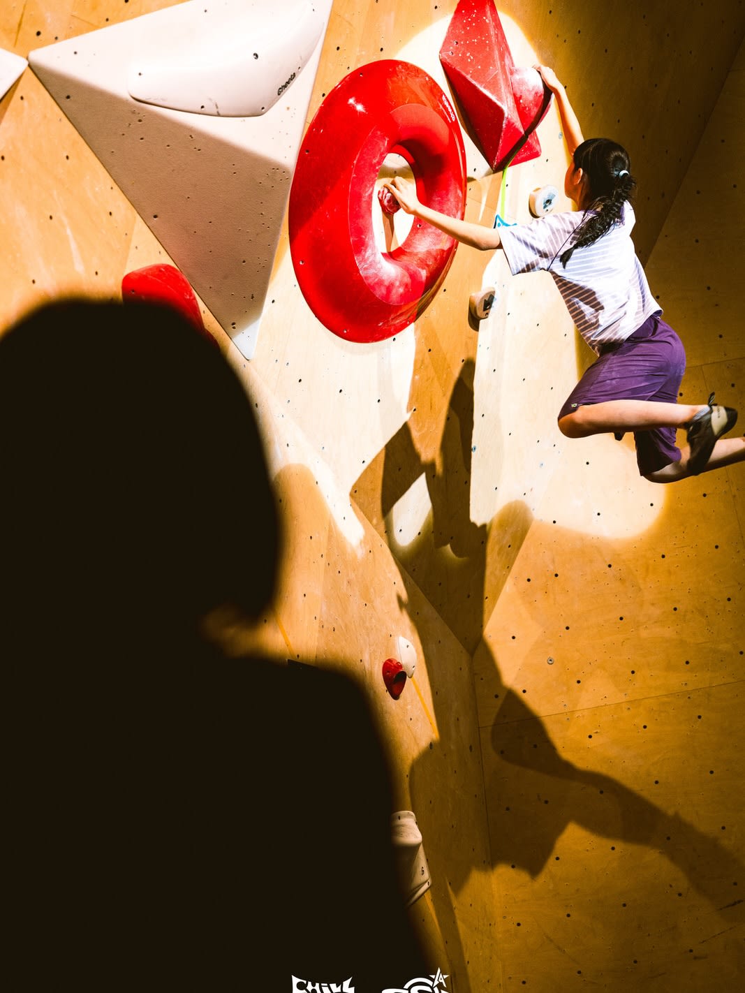 A dark silhouette of a person stands in the foreground, while a girl climbs a wooden wall with colorful holds in the background.