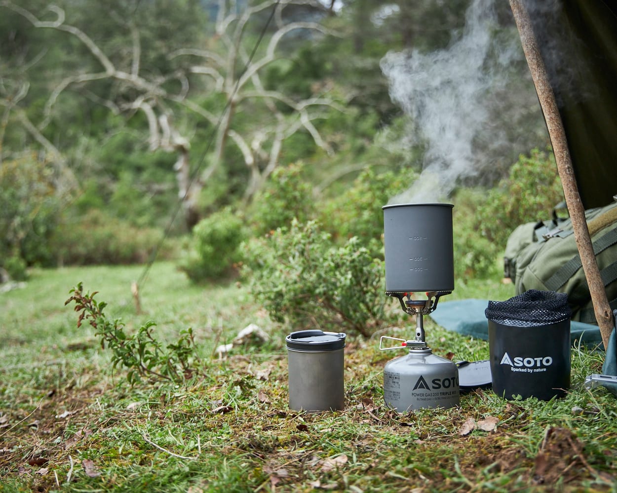 A camping stove with a steaming pot and a metal cup sit on a grassy forest floor in front of a tent and lush greenery.