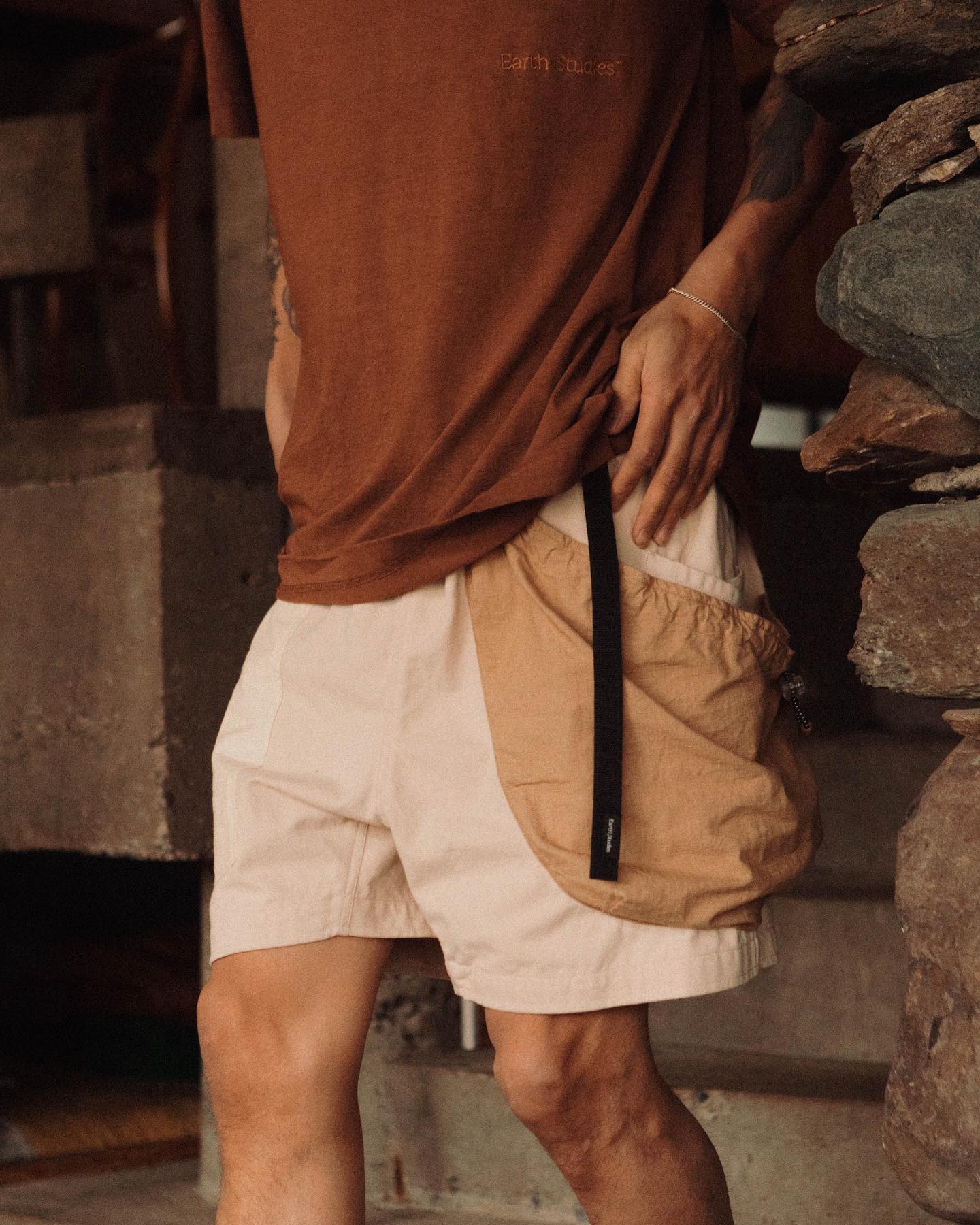 A person in a brown t-shirt and light-colored shorts with a tan pocket stands against a background of rough stone and concrete blocks.