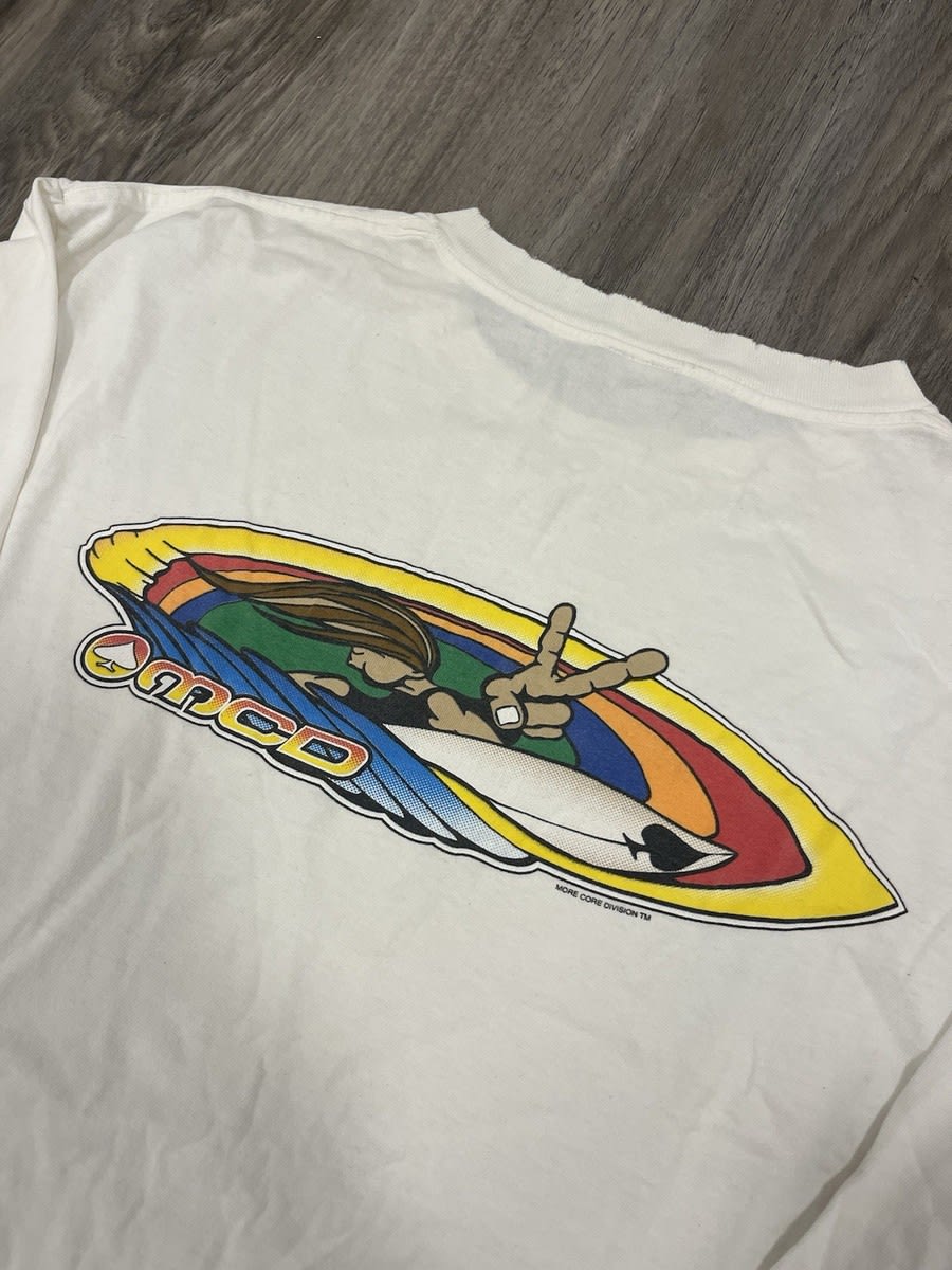 A colorful graphic of a surfer on a white board is printed on a white t-shirt resting on a grey wooden floor.