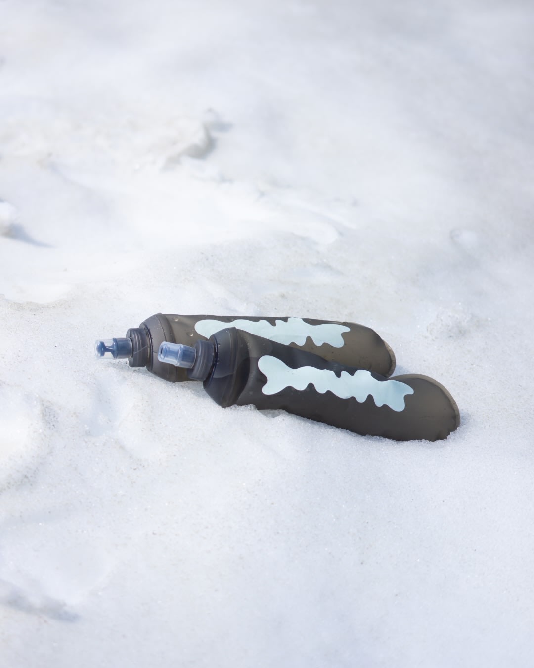Two grey soft flasks with white markings lie on a field of white snow.