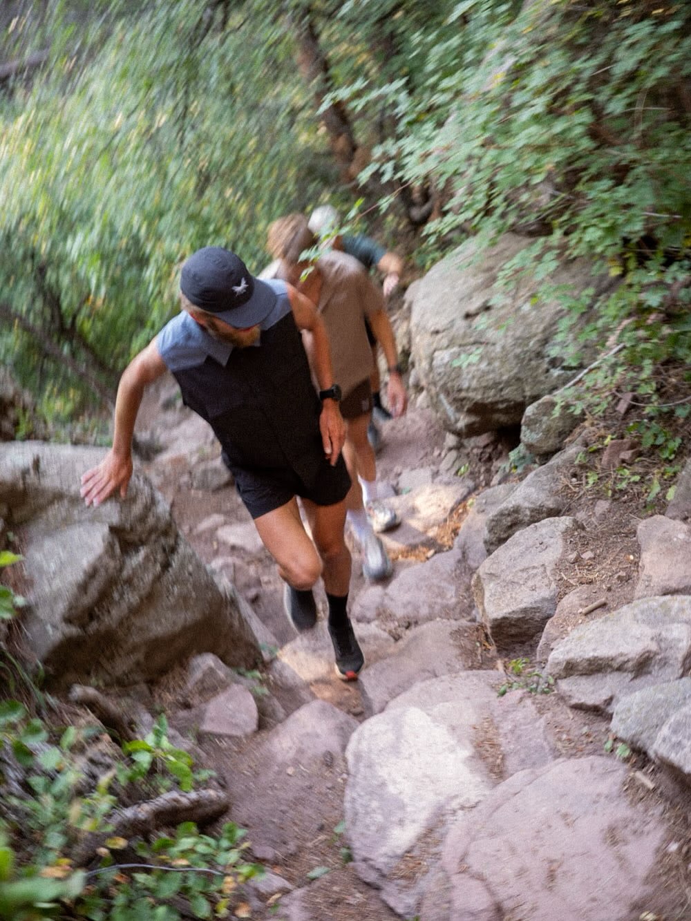Two hikers climb up a rocky, steep trail surrounded by dense green foliage.