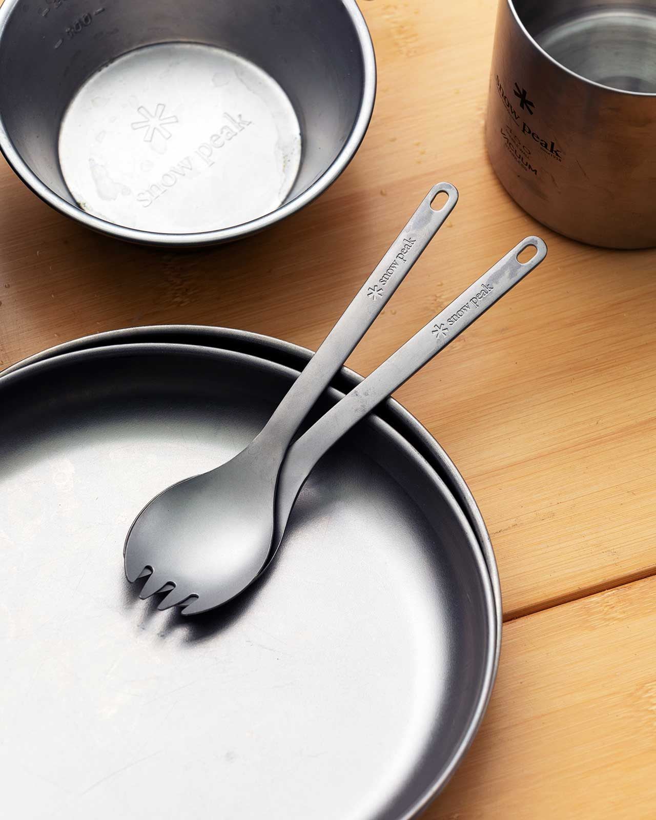 Two titanium sporks rest on a metal plate in the foreground, with a bowl and a cup sitting on a wooden surface in the background.
