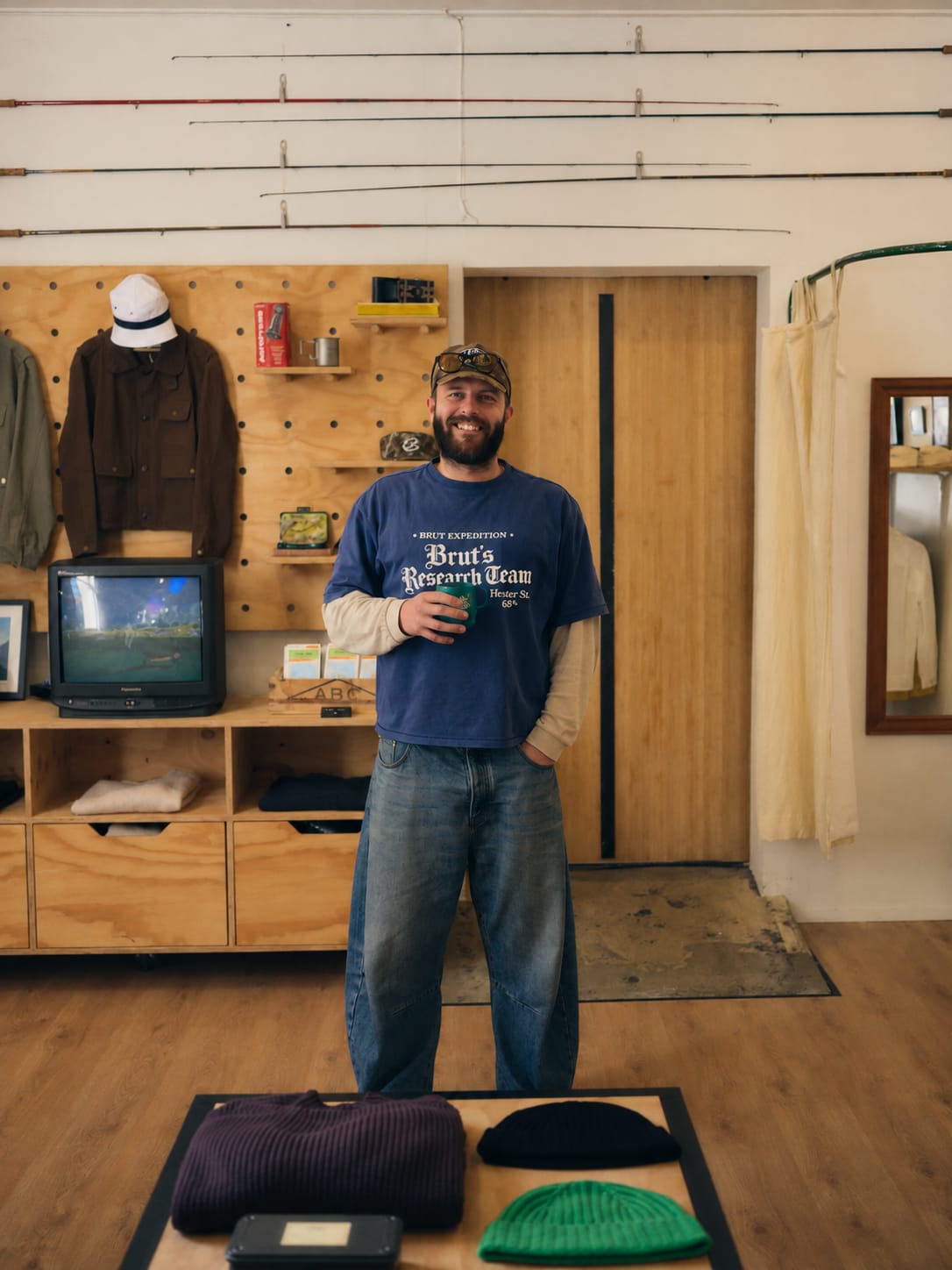 A smiling man in a blue t-shirt and jeans stands in the foreground of a rustic shop filled with outdoor gear and fishing rods.