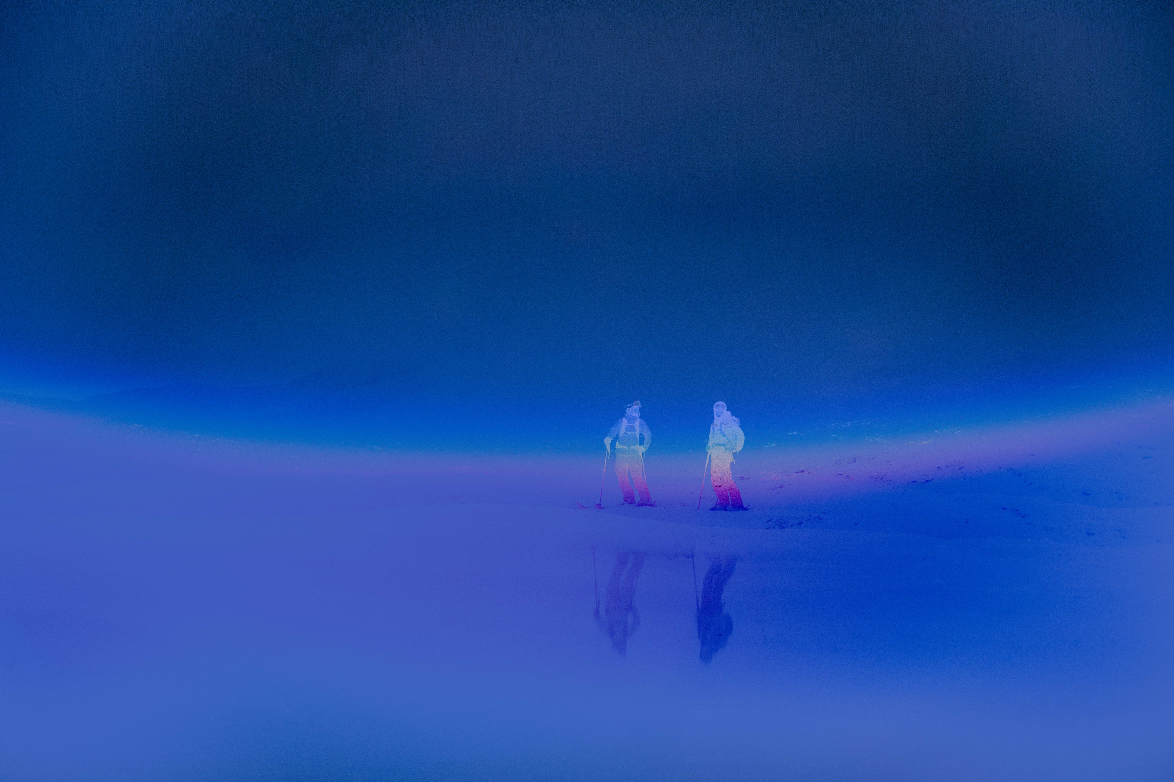 Two figures in ski gear stand on a reflective surface in the foreground, with a vast, dark blue expanse stretching into the background.