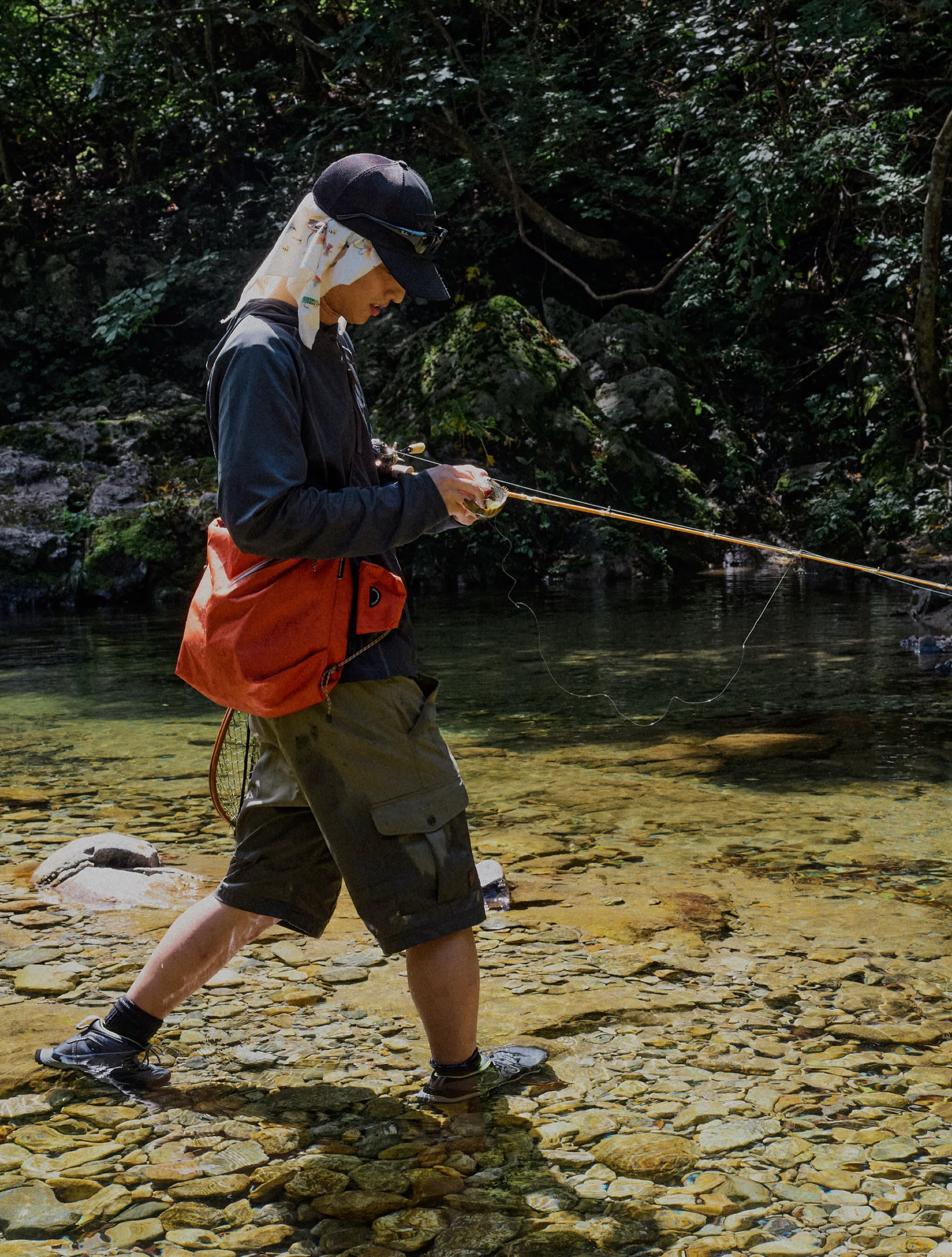 A man in fishing gear wades through a shallow, rocky stream against a backdrop of dense green foliage.