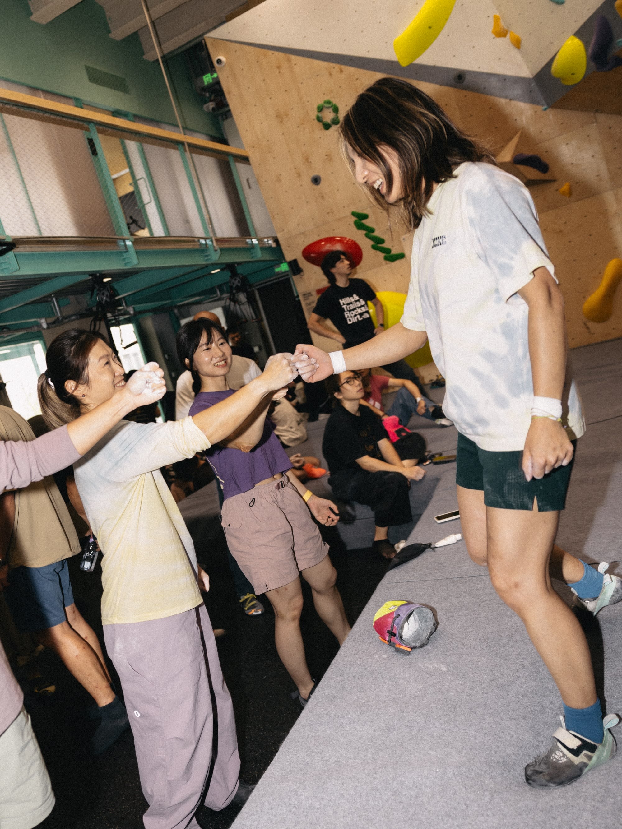 In the foreground, two women fist-bump on a climbing wall, while in the background, other people watch and relax in a brightly lit gym.