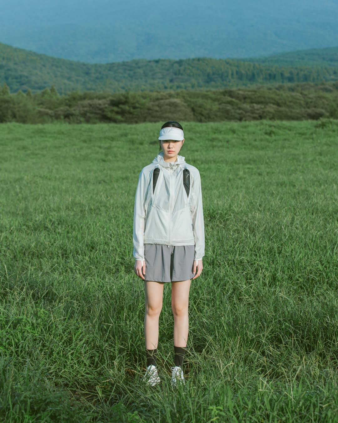 A woman in athletic wear stands in a lush green field with rolling forested hills in the background.