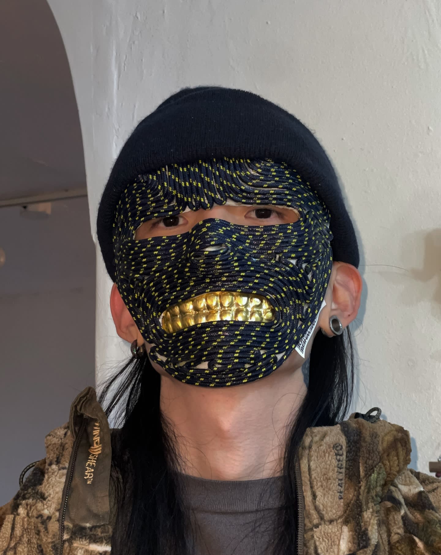 A person wearing a black beanie, a woven black and yellow mask with gold teeth, and a camouflage jacket stands against a plain white wall.