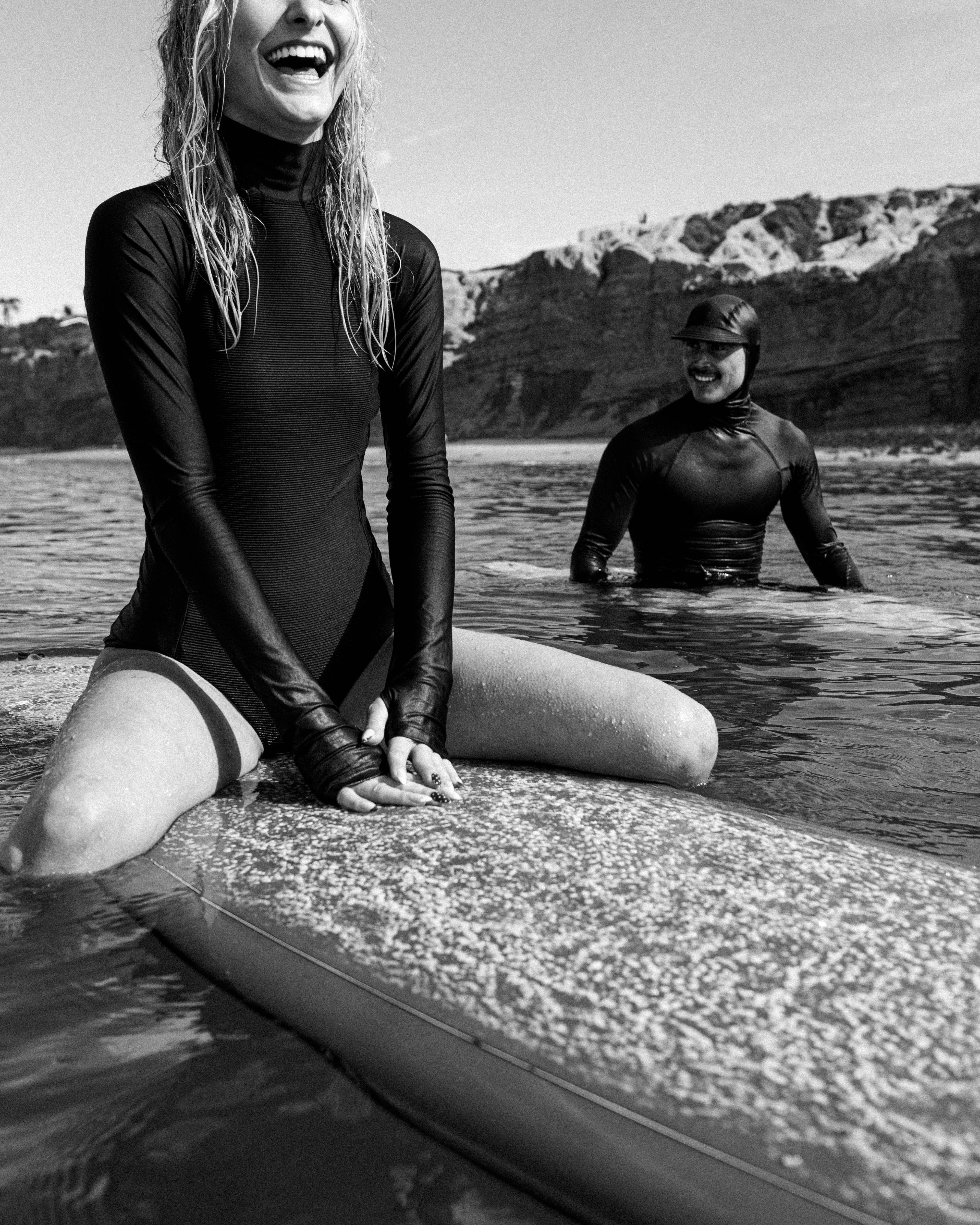 A laughing woman in a black wetsuit sits on a surfboard in the foreground, while a man in a wetsuit stands in the water against a backdrop of rugged cliffs.