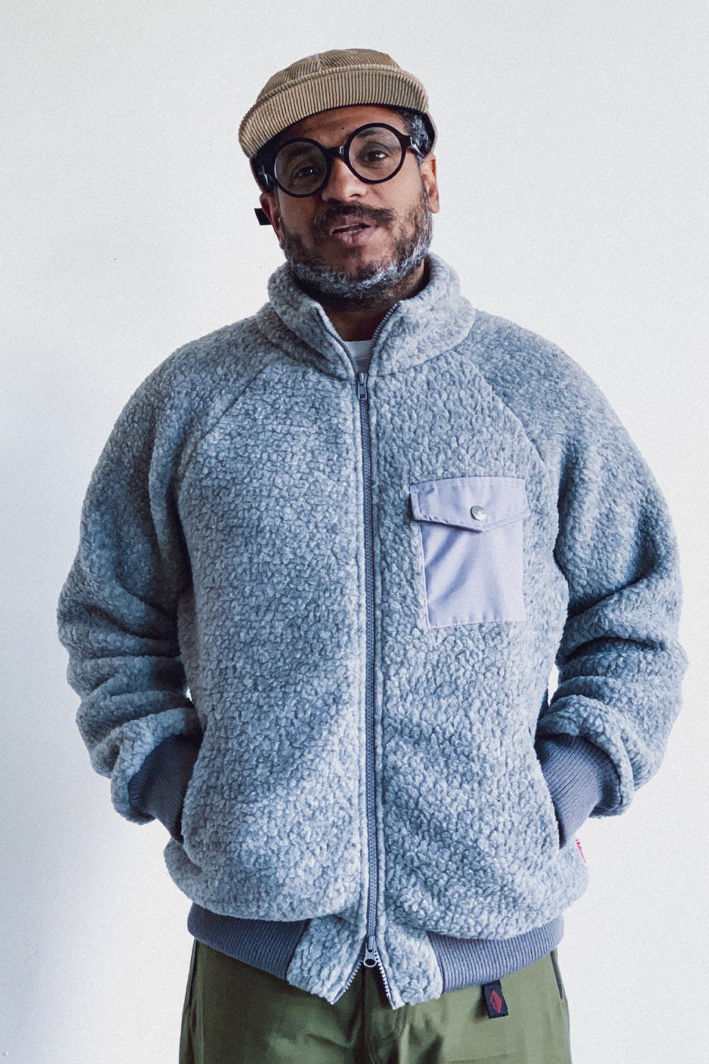 A man in a fuzzy gray jacket and a cap stands against a plain white background.