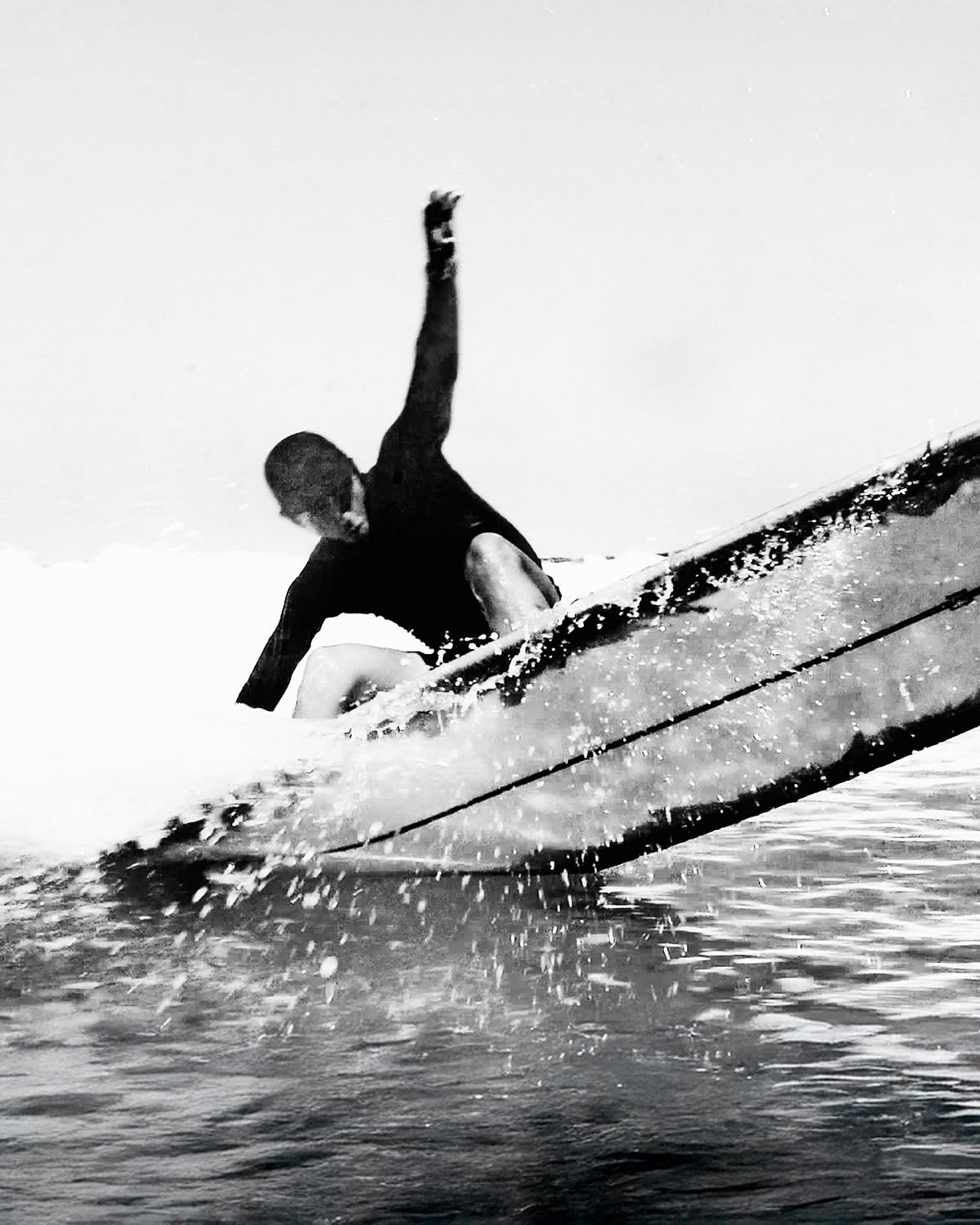 A surfer in a dark wetsuit rides a breaking wave in the foreground against a pale, clear sky in the background.