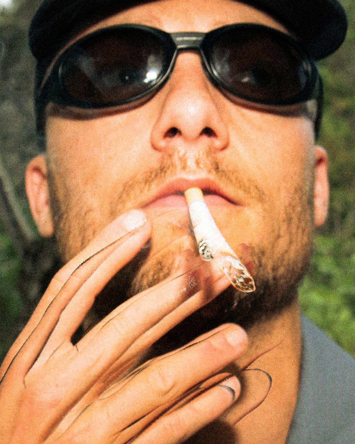 A man wearing sunglasses and a black hat smokes a joint in the foreground against a blurred green natural background.