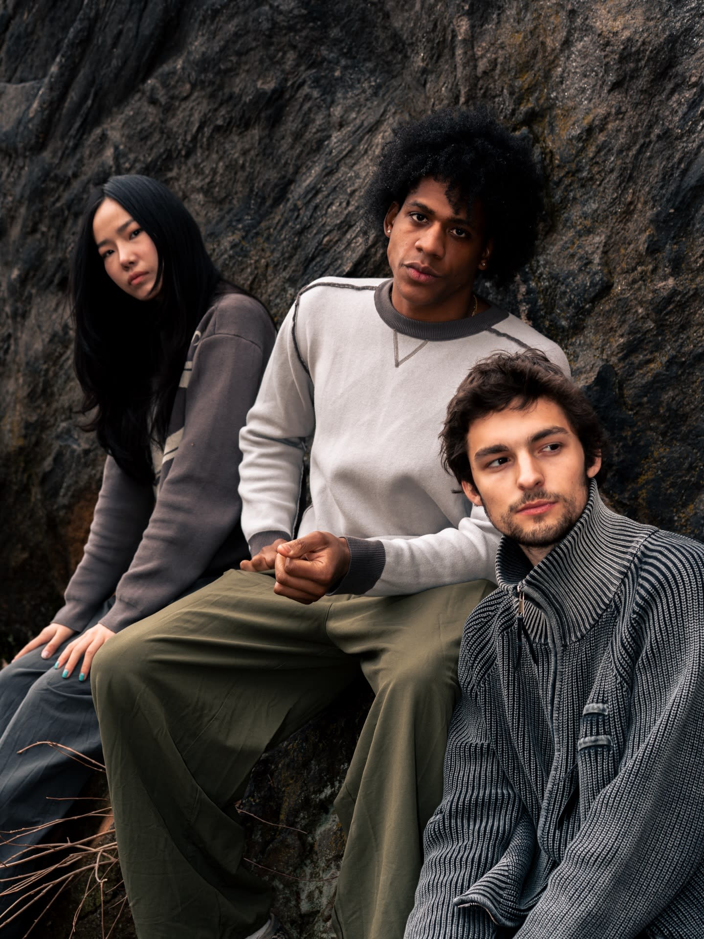Three people wearing neutral-toned sweaters pose against a dark, textured rock wall.
