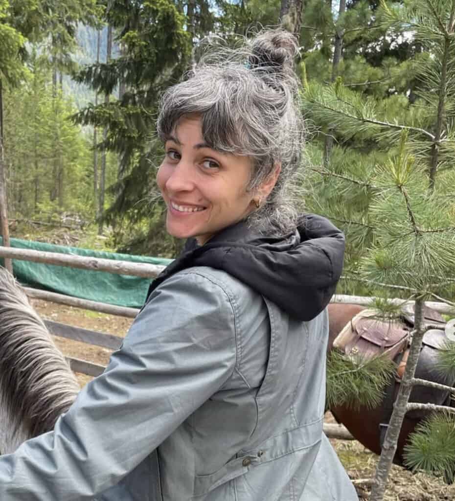 A smiling woman with graying hair pulled up in a bun, wearing a gray jacket, sits on a horse in the foreground, with a wooded area and a fence in the background.