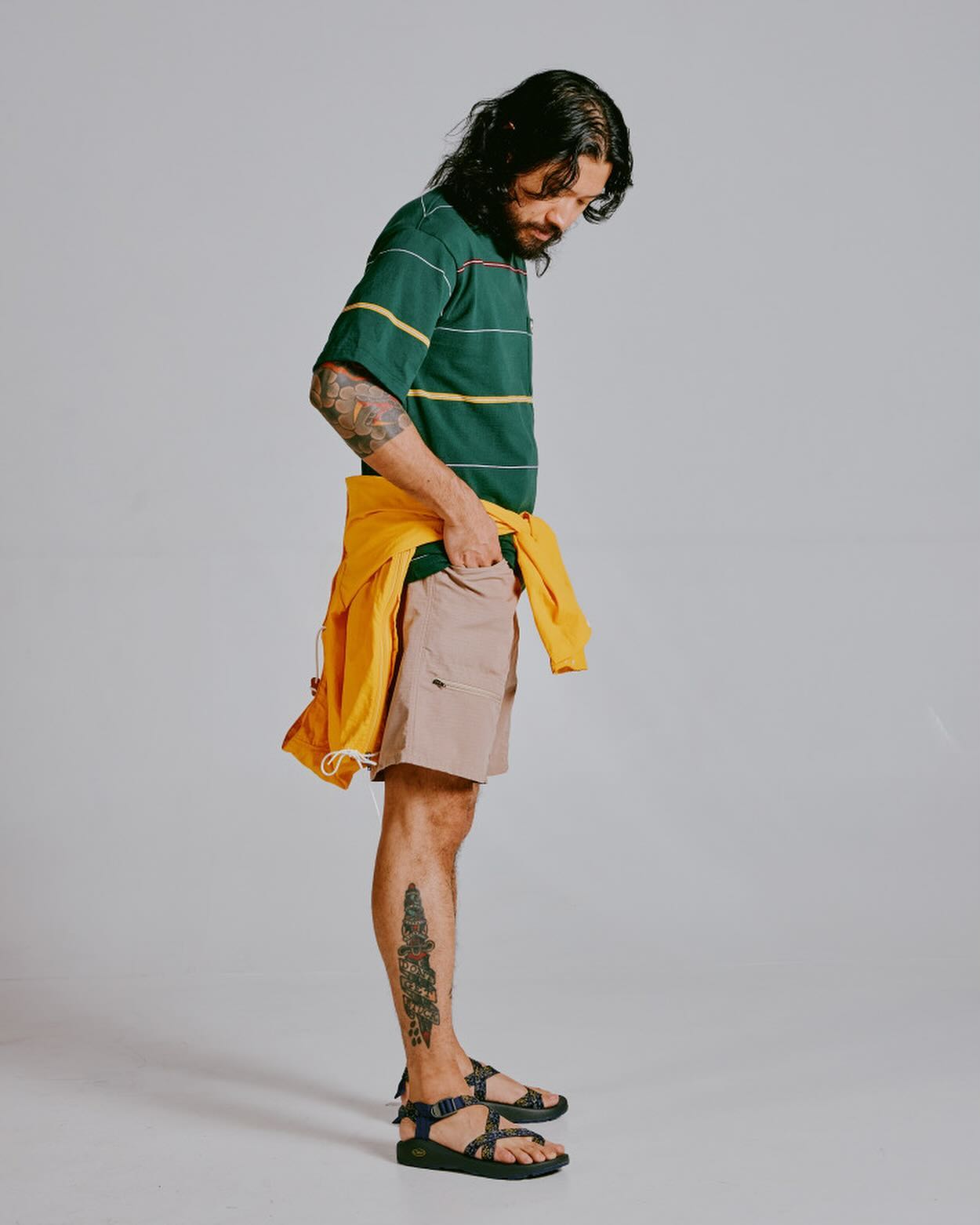 A man with long dark hair and tattoos stands in profile, wearing a green and yellow striped shirt, tan shorts, and black sandals, with a yellow sweatshirt tied around his waist, against a plain gray background.