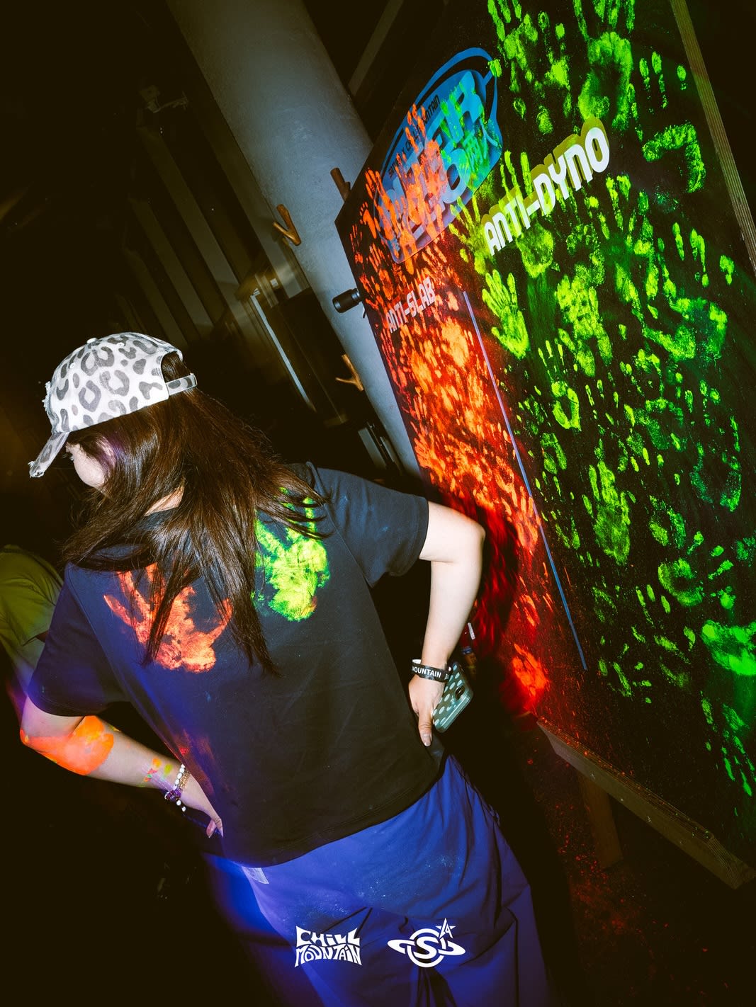 A woman in a leopard-print cap and black t-shirt stands in the foreground before a dark wall covered in glowing neon green and orange paint splatters.