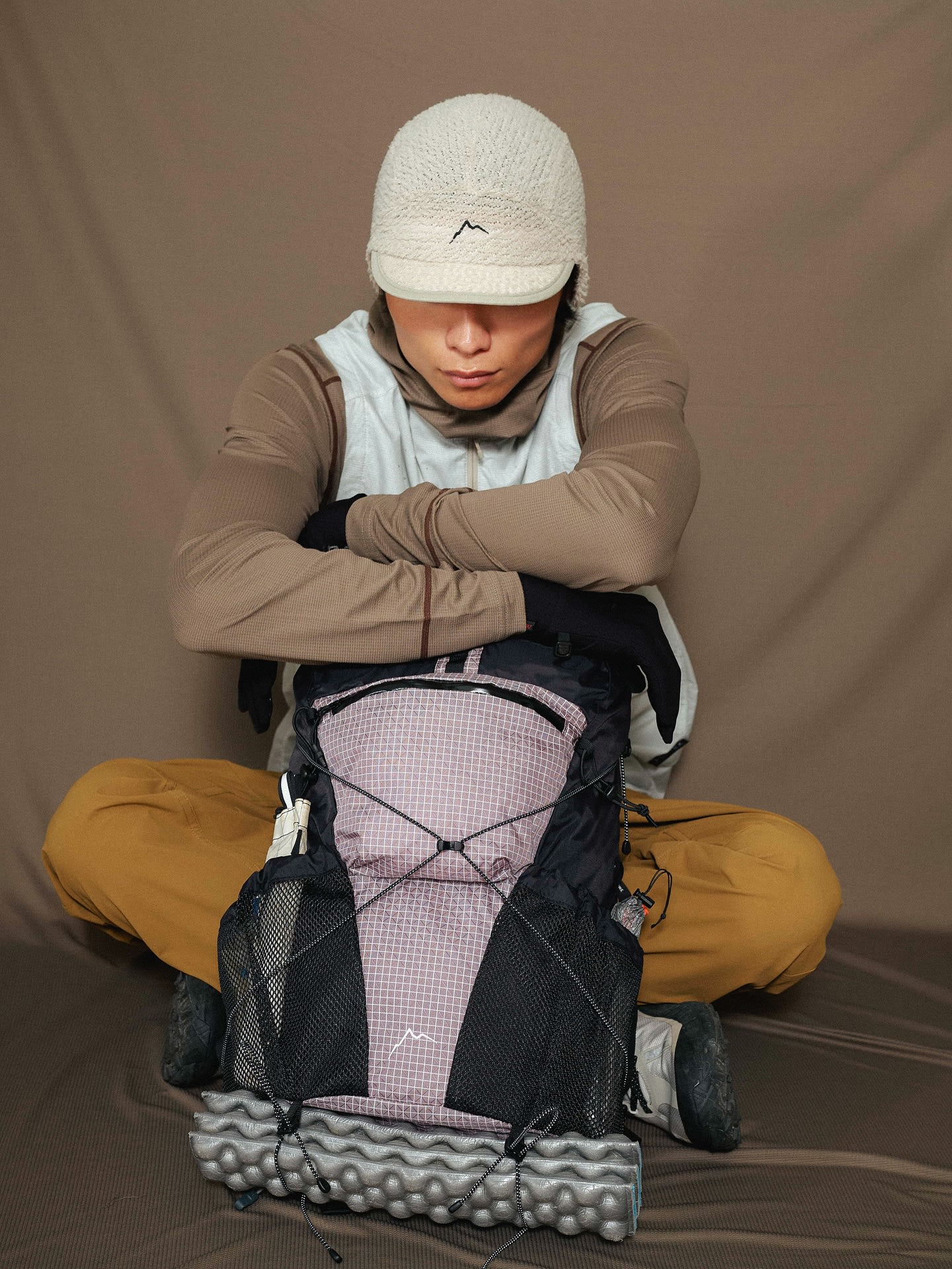 A man in outdoor gear sits cross-legged with his arms resting on a backpack against a plain brown backdrop.