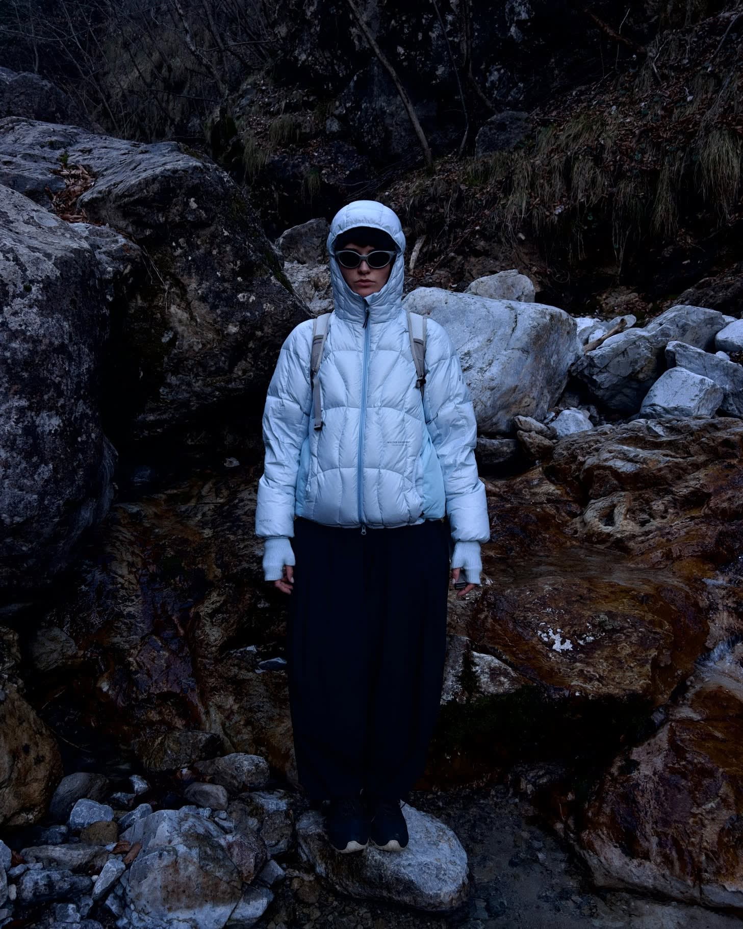 A person wearing a light blue hooded puffer jacket and dark pants stands on a rock in a rugged, rocky landscape.