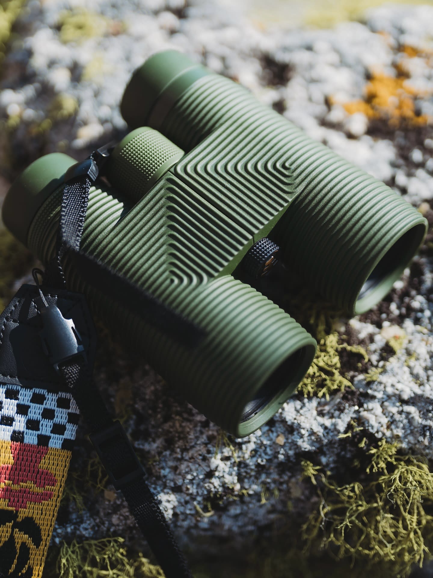 Green binoculars with a patterned strap rest on a moss-covered rock, with a blurred, natural background.