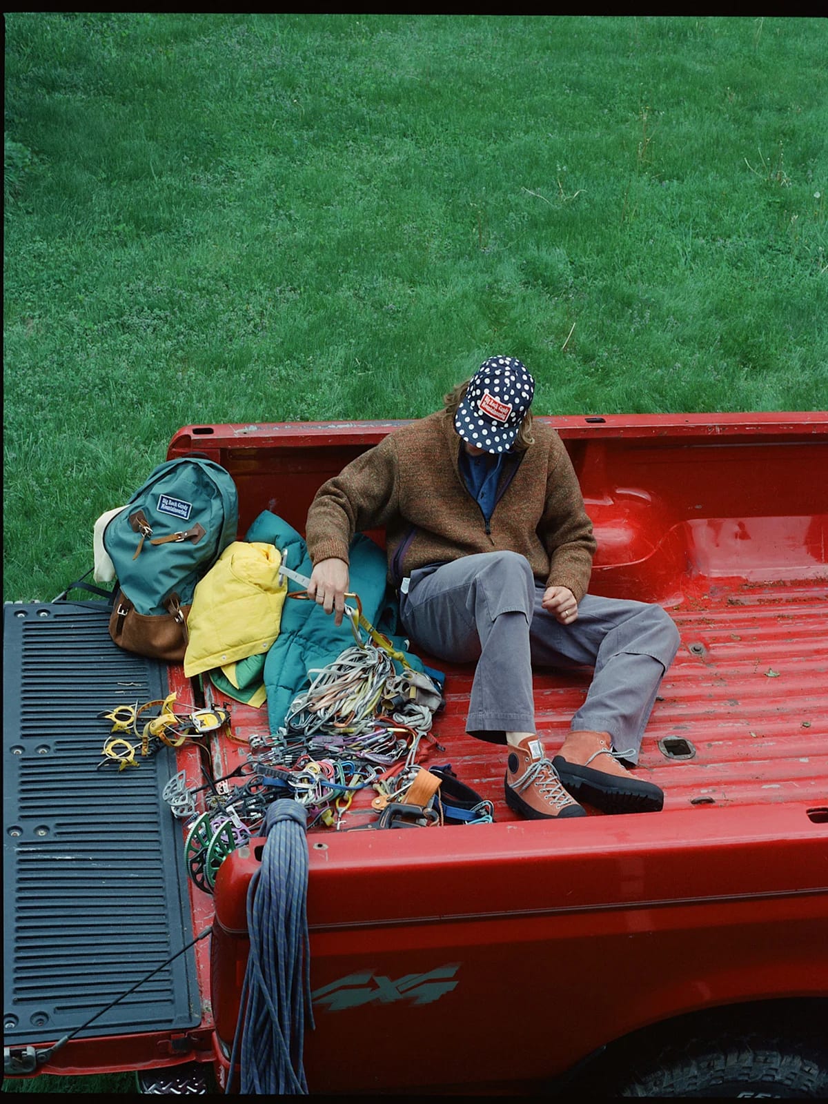 A person wearing an American flag hat sits in the bed of a red pickup truck filled with climbing gear, with a lush green lawn in the background.
