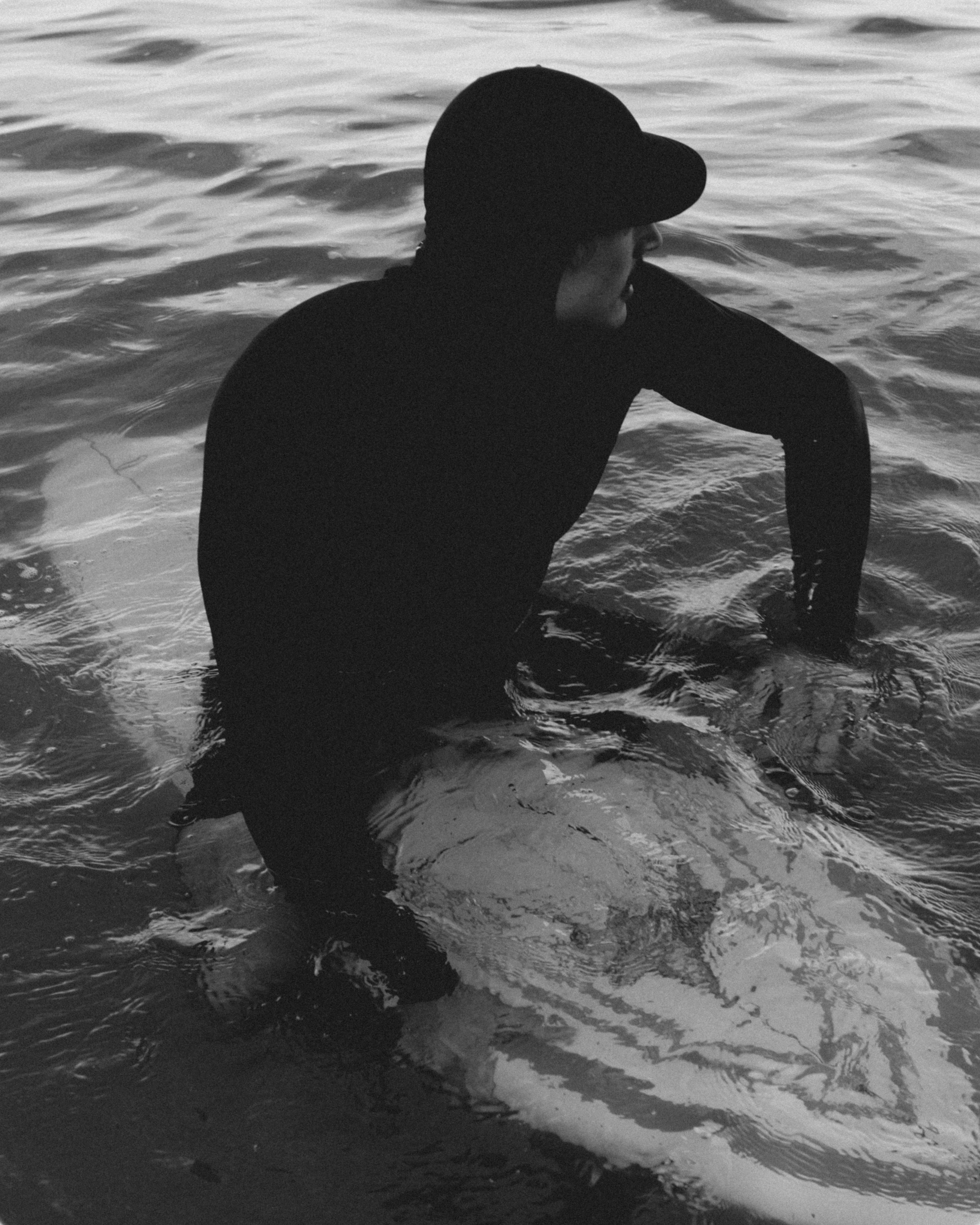 A person in a black wetsuit and cap sits on a surfboard amidst rippling ocean water.
