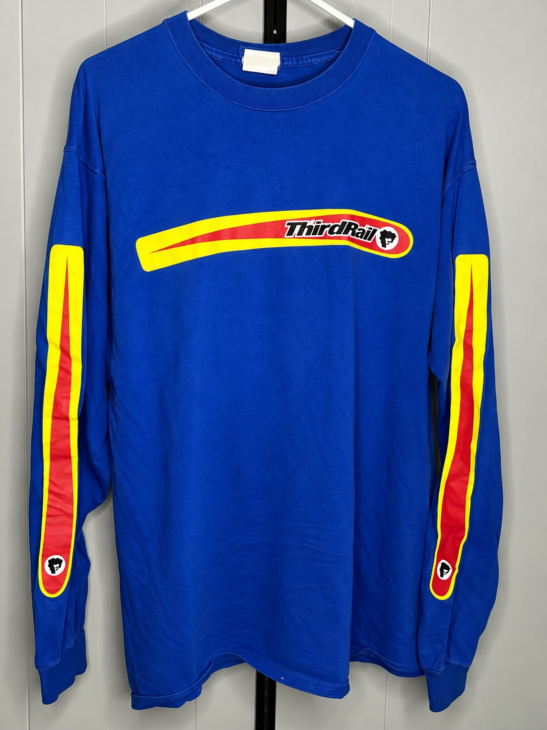 A blue long-sleeved shirt with yellow and red "Third Rail" branding is displayed on a hanger against a plain grey wall.