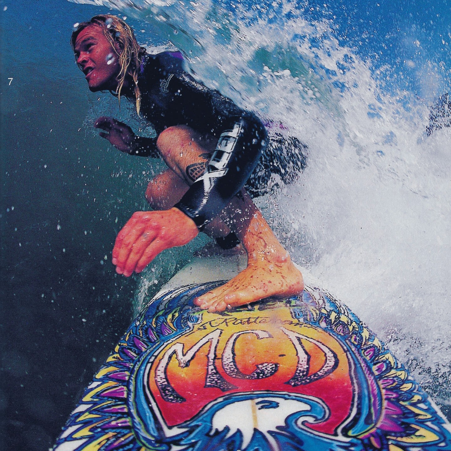A surfer in a black wetsuit rides a colorful surfboard through a crashing white wave under a clear blue sky.