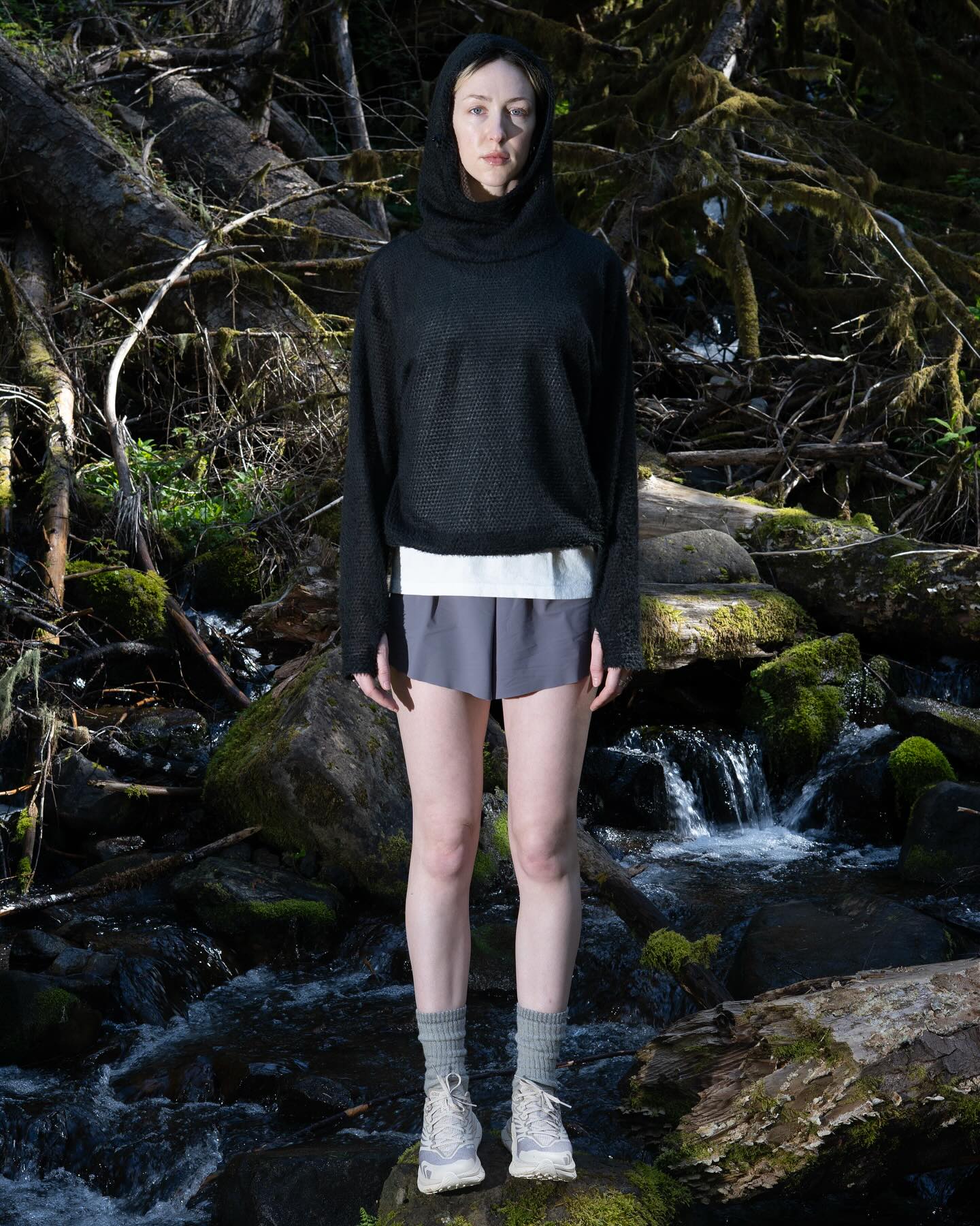 A woman wearing a black hooded top and grey shorts stands on mossy rocks in front of a flowing stream and a dense, wooded background.