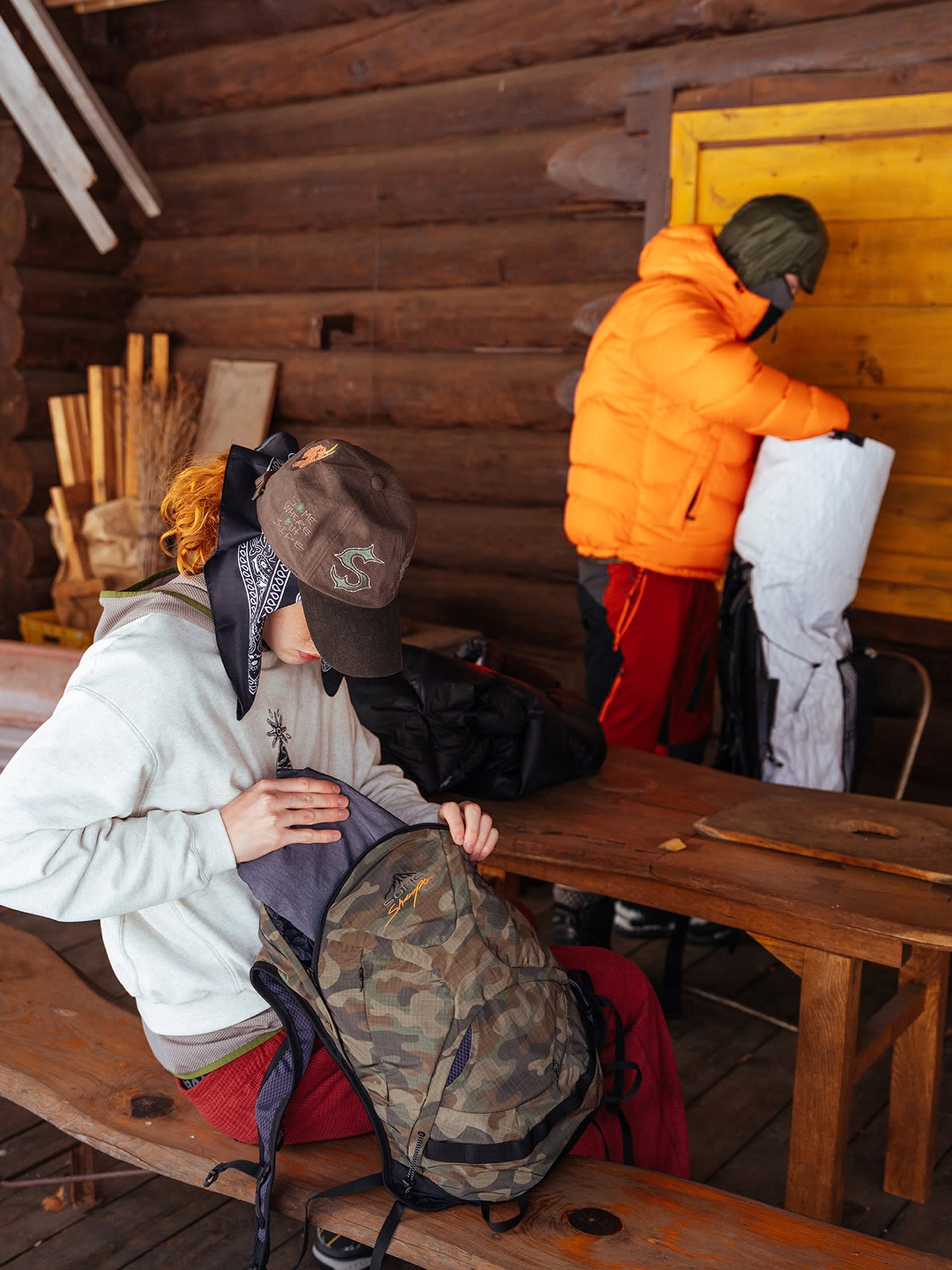 A person in a white sweatshirt and brown cap packs a camouflage backpack in the foreground, while another person in an orange jacket stands by a yellow door in the background of a rustic wooden cabin.