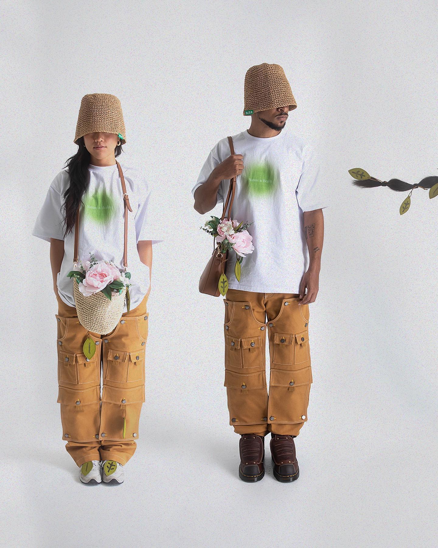 Two people in bucket hats and cargo pants stand against a white background, with a branch of leaves visible on the right.