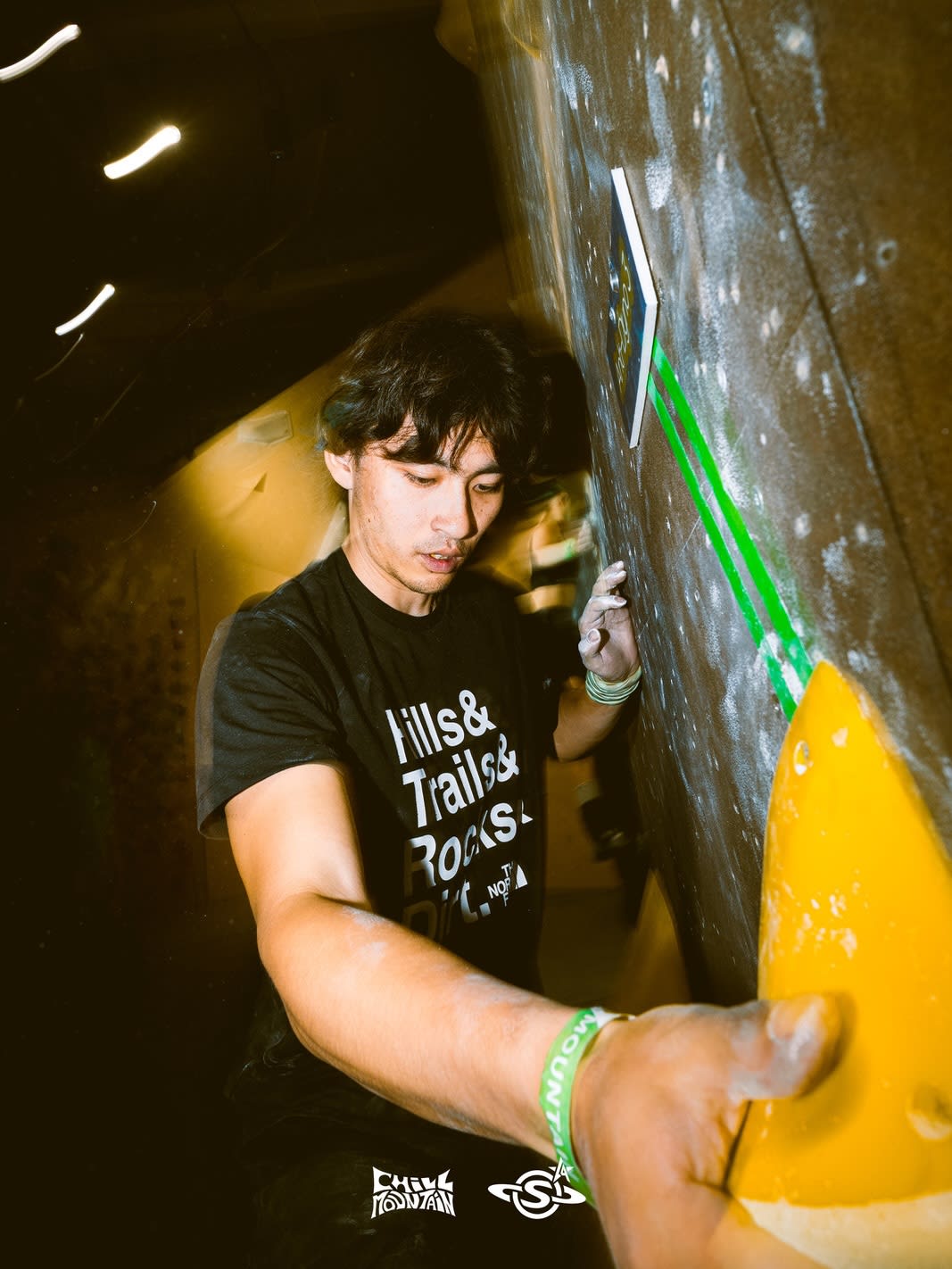 A man in a black t-shirt climbs a dark indoor wall, gripping a bright yellow hold against a dimly lit background.