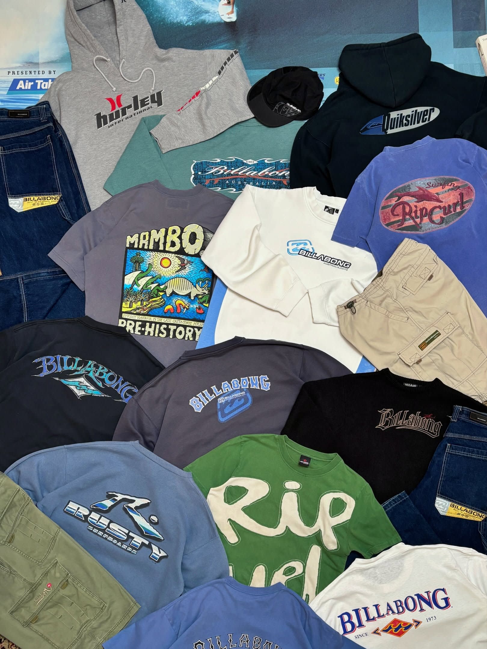 A variety of surf-brand hoodies, t-shirts, and pants are spread across a blue surface.