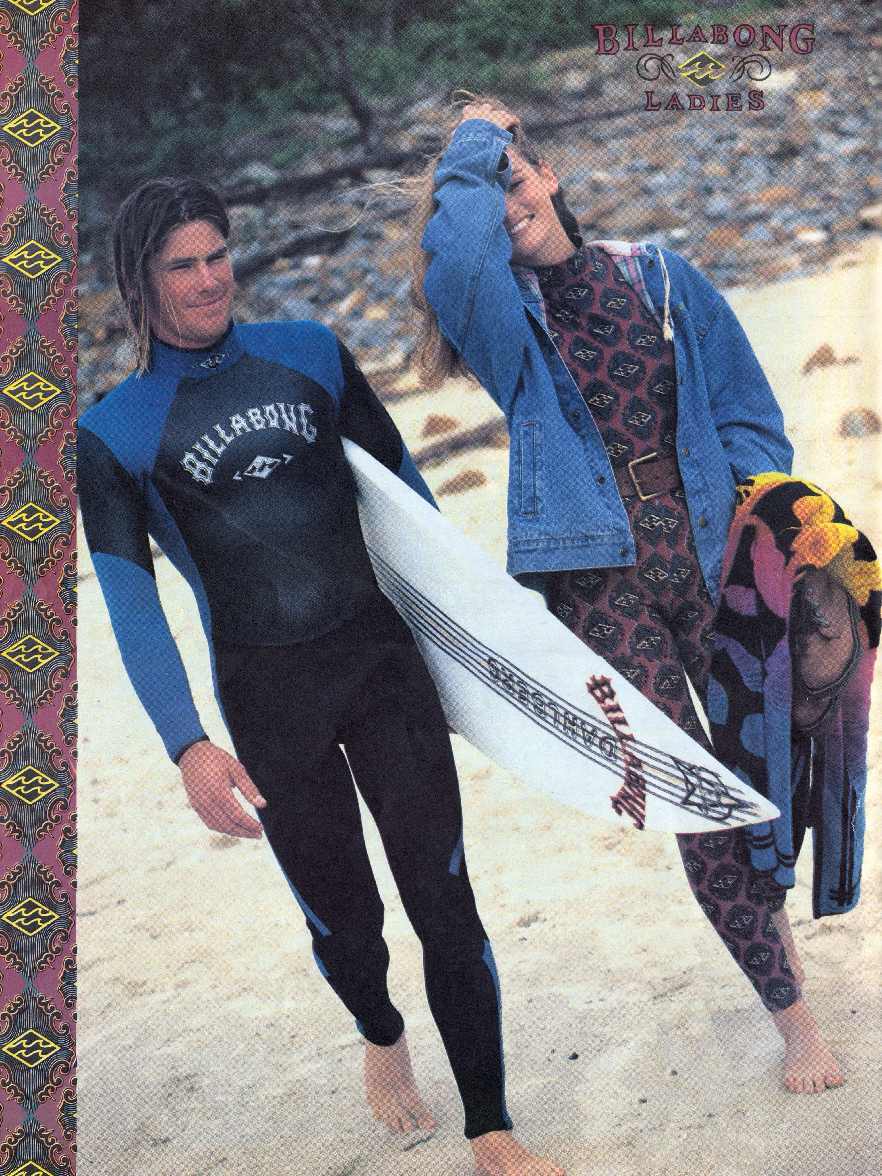 A man in a blue and black wetsuit holding a surfboard stands beside a woman in a denim jacket and patterned bodysuit on a sandy beach with greenery in the background.
