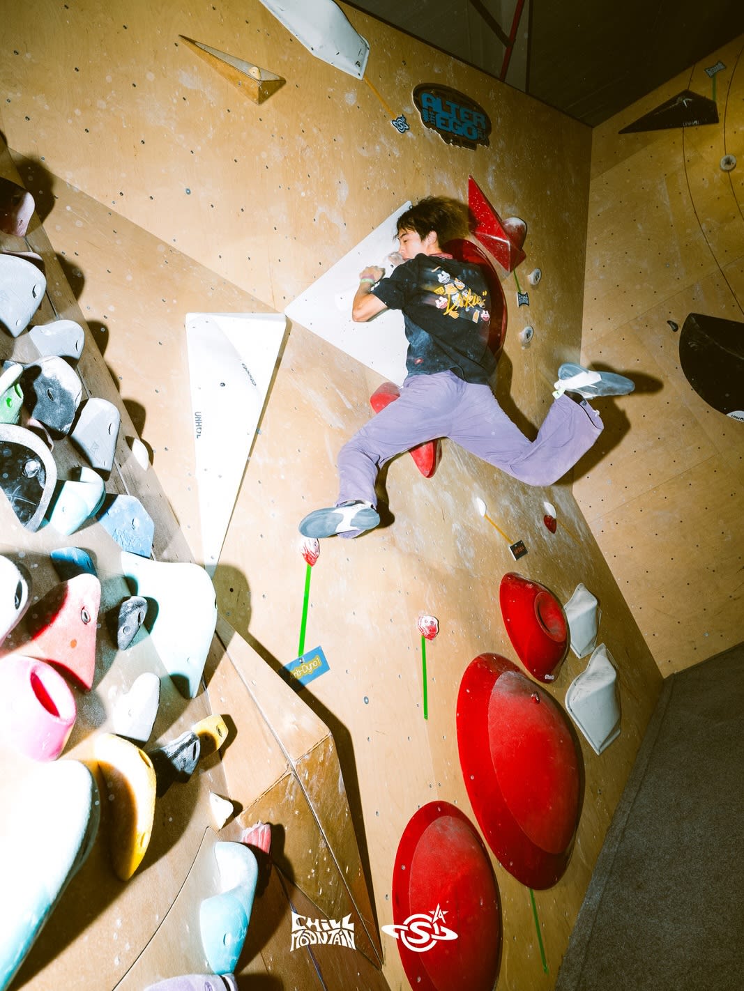 A climber in a black t-shirt and purple pants scales a wooden bouldering wall equipped with various colorful holds.