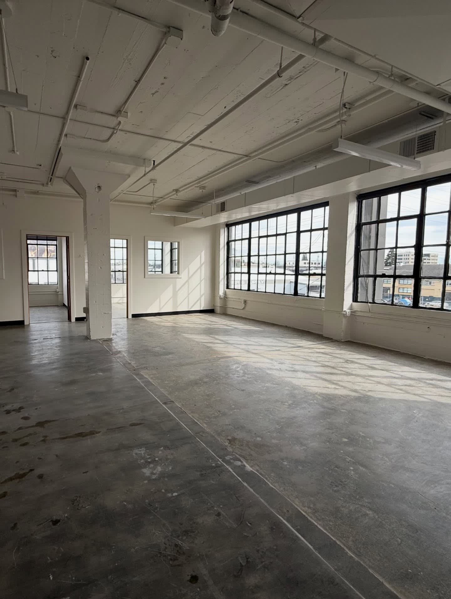 An empty industrial room with a concrete floor in the foreground and large grid windows overlooking a city in the background.