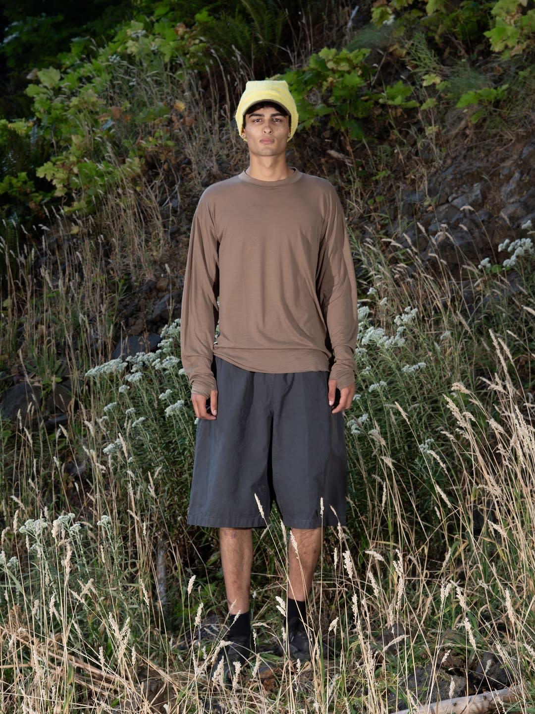 A man wearing a yellow beanie, brown long-sleeved shirt, and grey shorts stands in a field of tall grass against a backdrop of rocky terrain and green foliage.