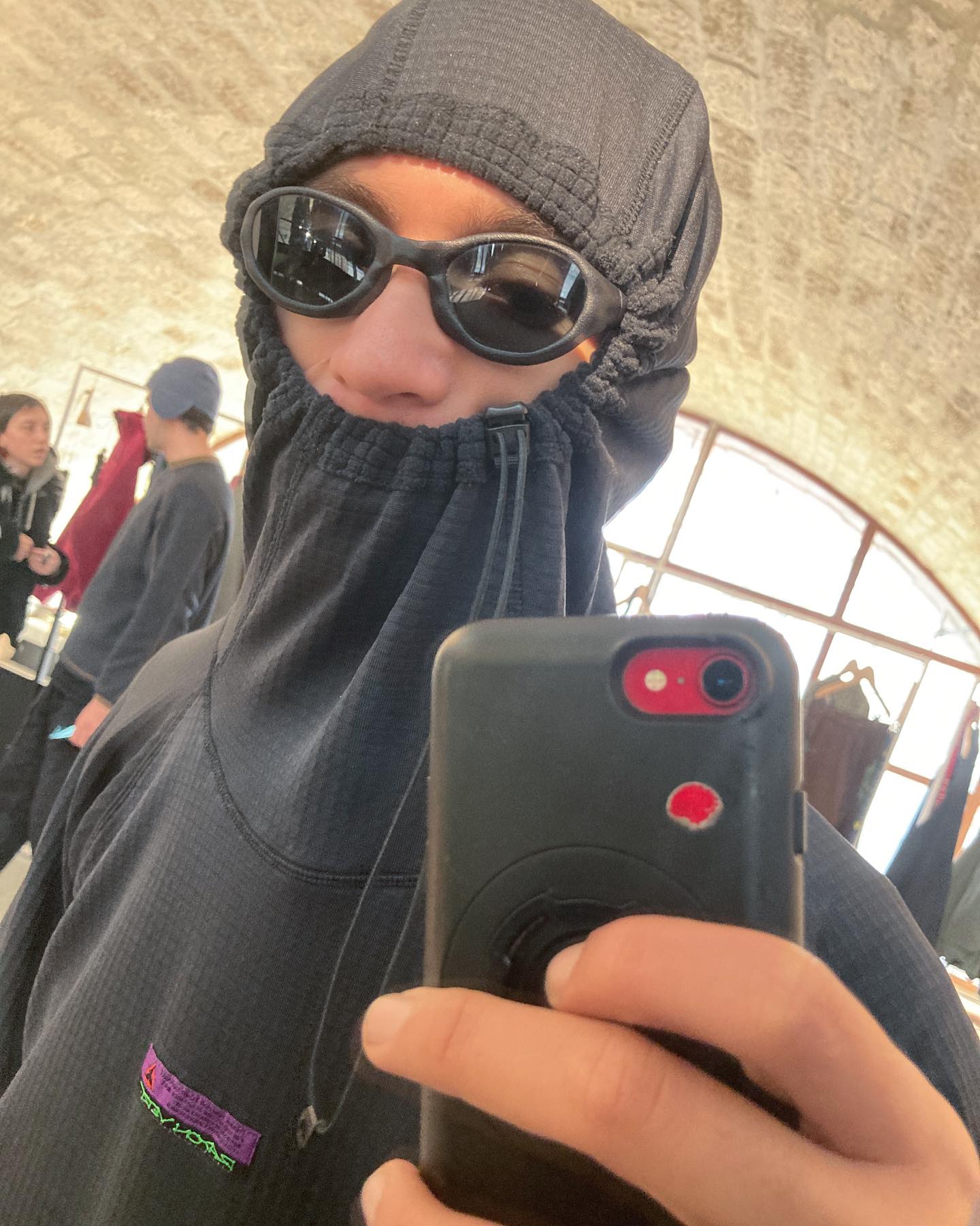 A person wearing a black hooded face mask and sunglasses takes a mirror selfie in a room with a stone wall and other people in the background.