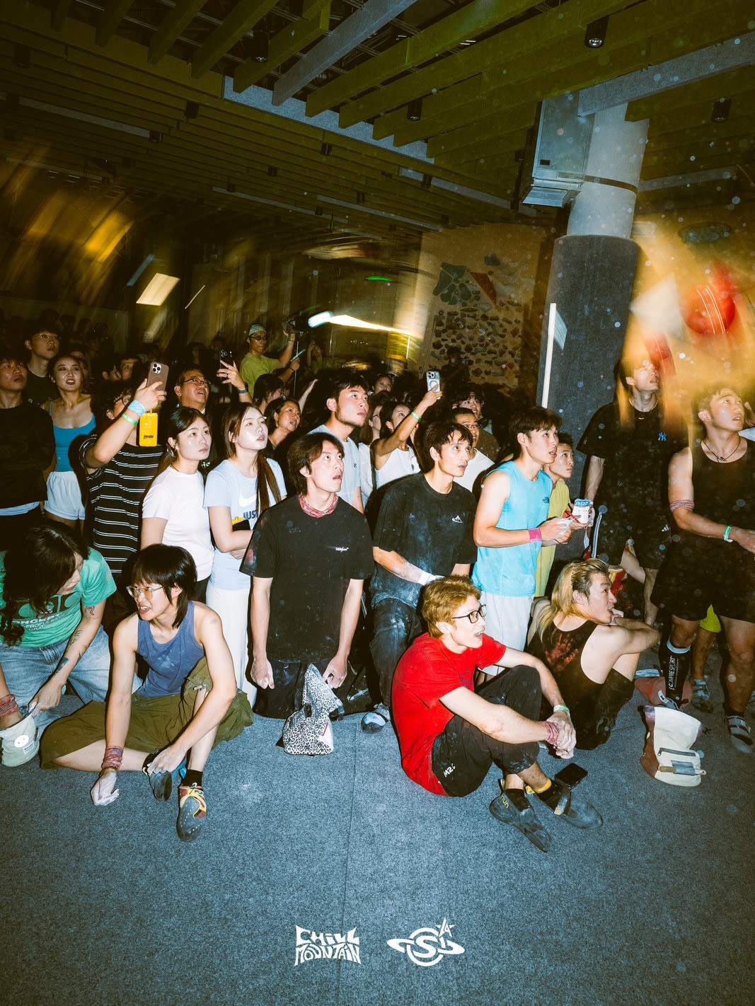 A crowd of young people gathers in a dimly lit indoor space with a beamed ceiling, some sitting on the floor in the foreground while others stand and record with phones in the background.