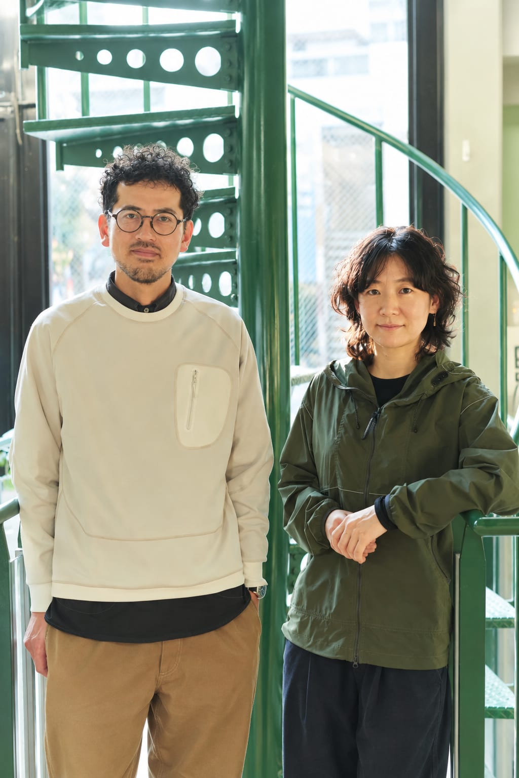 A man in a cream sweater and a woman in an olive jacket stand together in the foreground against the backdrop of a green spiral staircase.